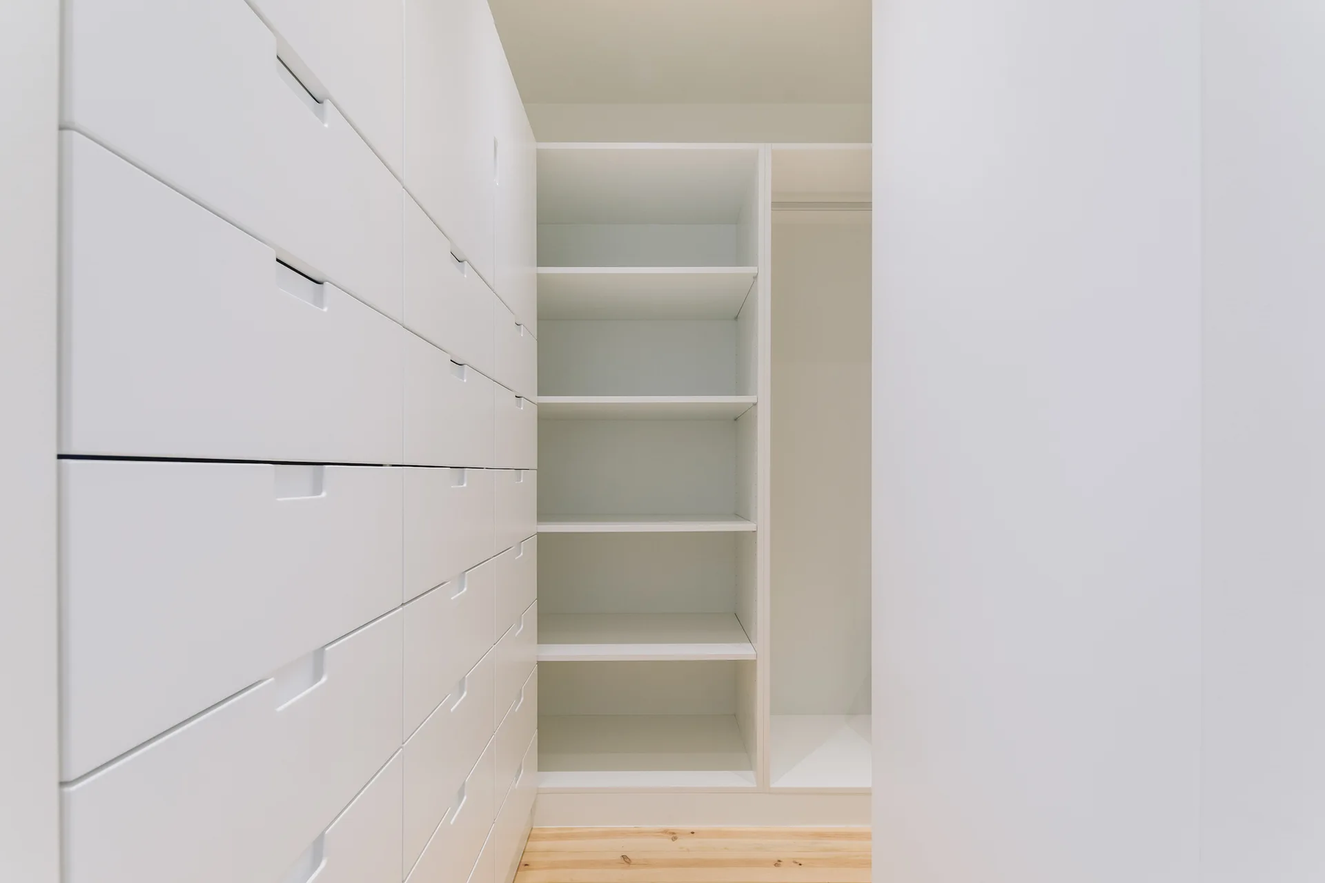 A minimalist white built-in closet features multiple rows of drawers with integrated handles, open shelving, and light wood flooring.