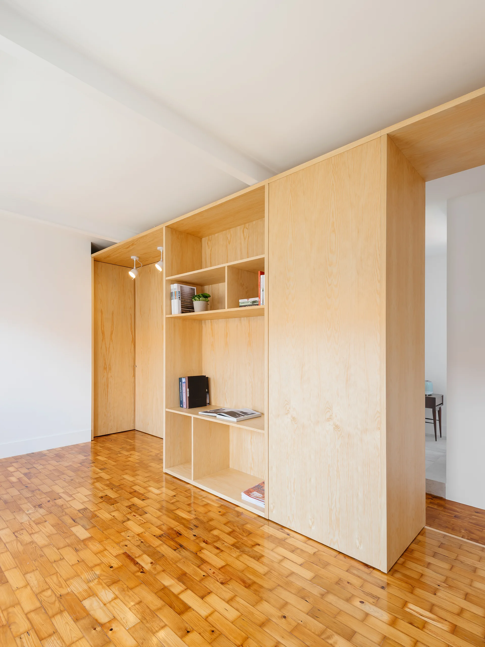 A custom light wood partition wall with integrated shelving stands on warm parquet flooring in a bright room.