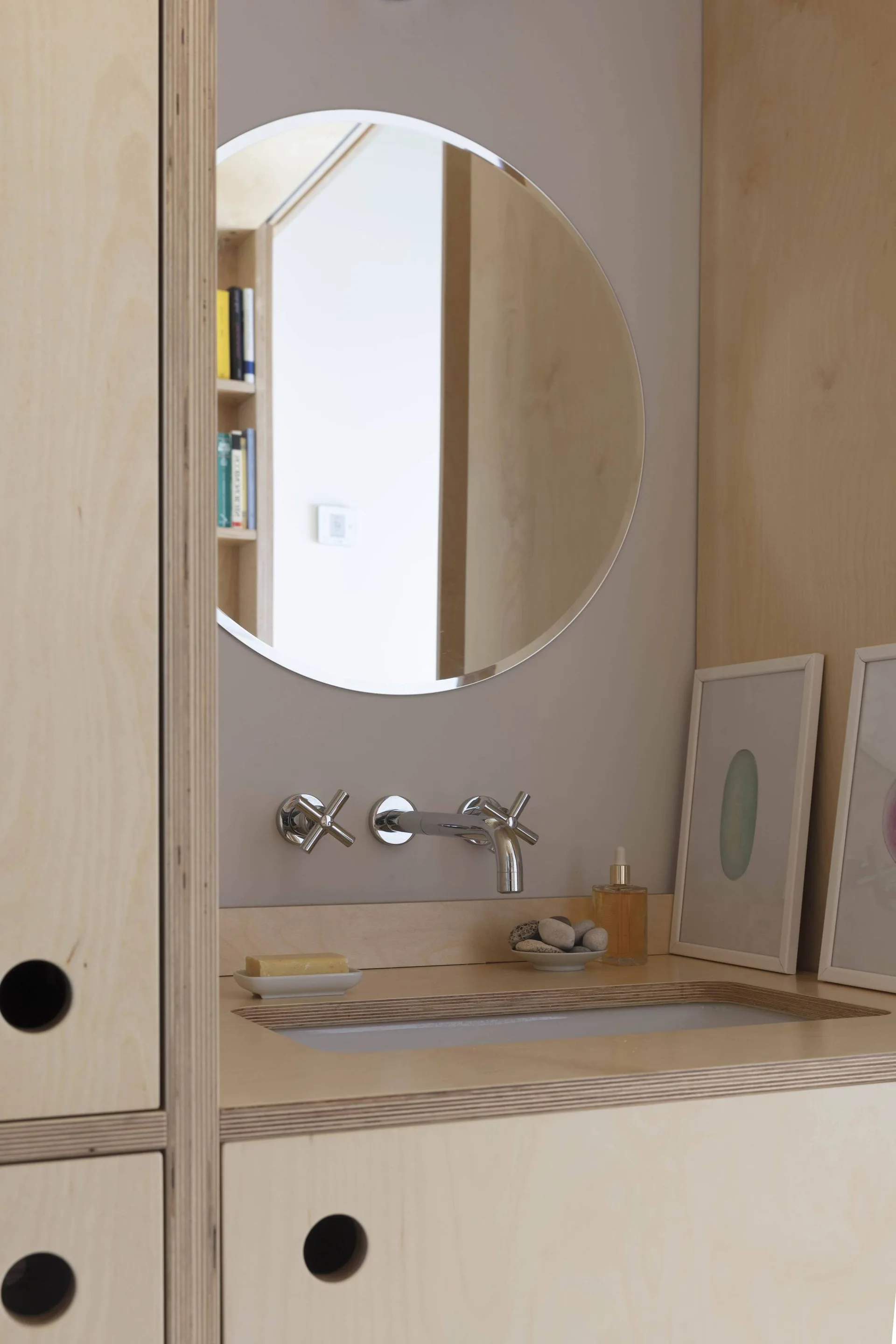 A round mirror and wall-mounted chrome faucet sit above a plywood vanity with an integrated sink and circular cabinet cutouts.