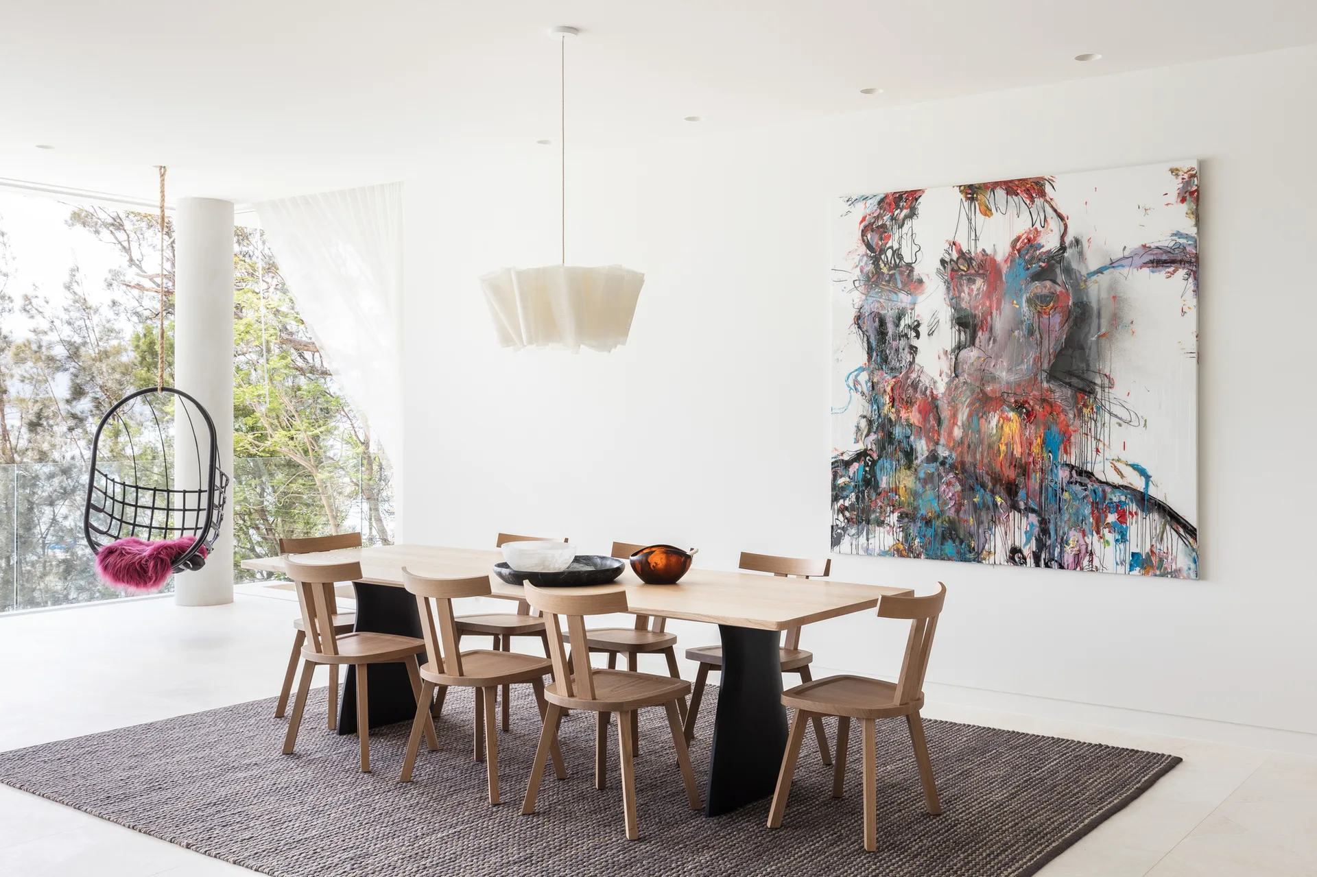 A minimalist dining room features a light wood table with black pedestals, matching chairs on a textured grey rug, and a large colorful abstract painting.