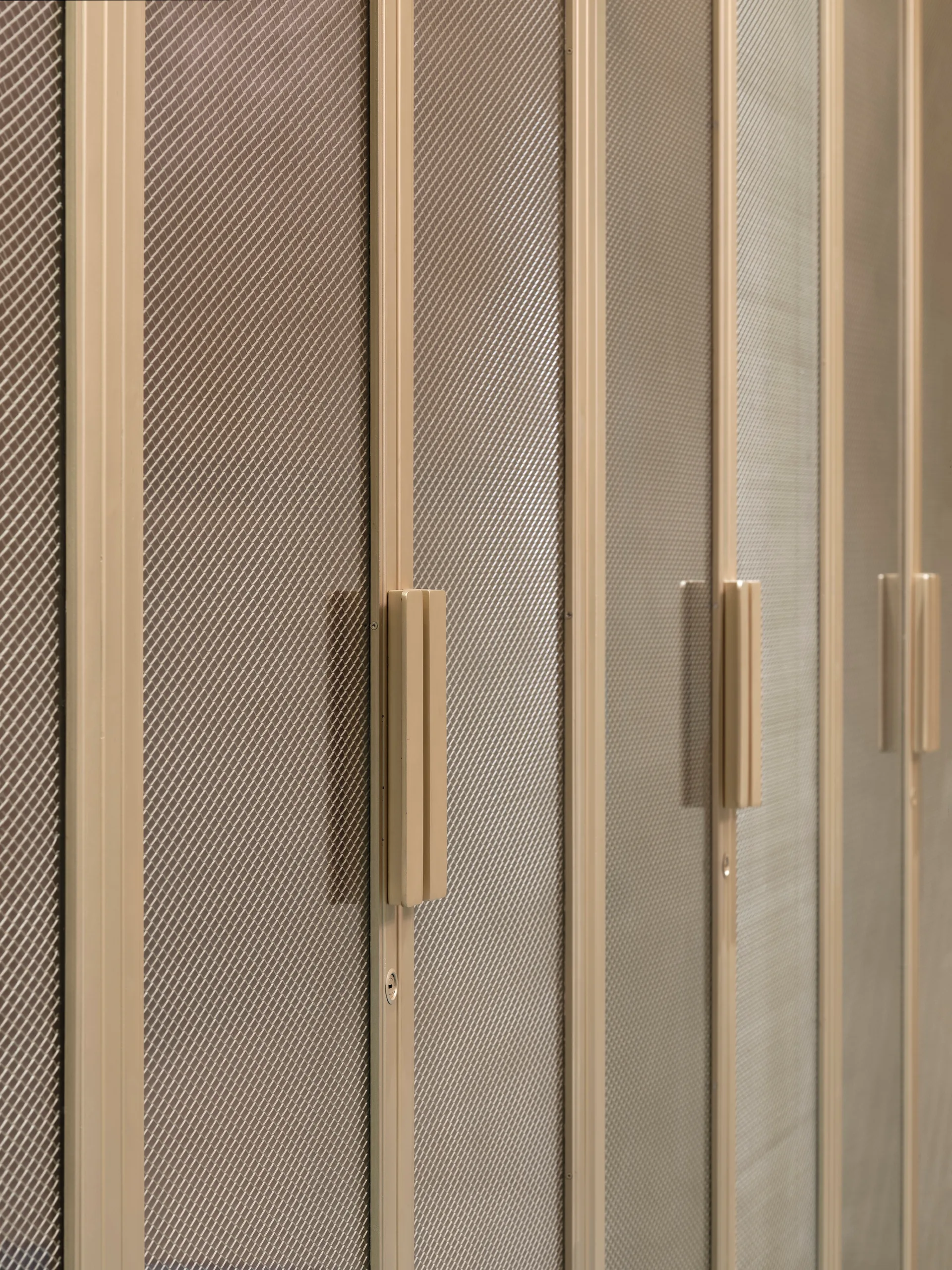 Close-up of beige metal wardrobe doors featuring diamond-patterned mesh panels and long vertical bar handles.