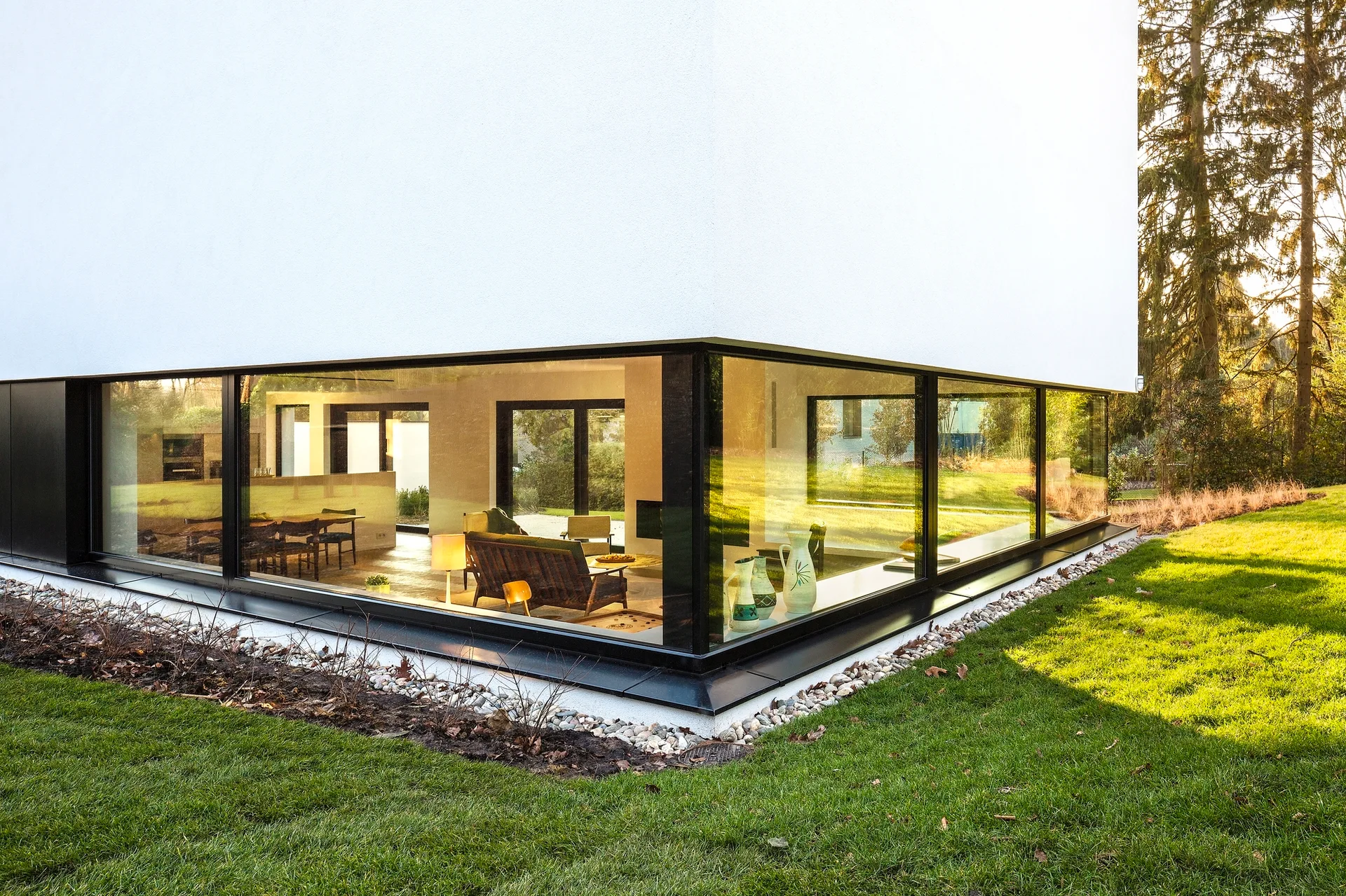 A modern white house features a large black-framed corner glass window showing an interior living room with a wood sofa and dining set.
