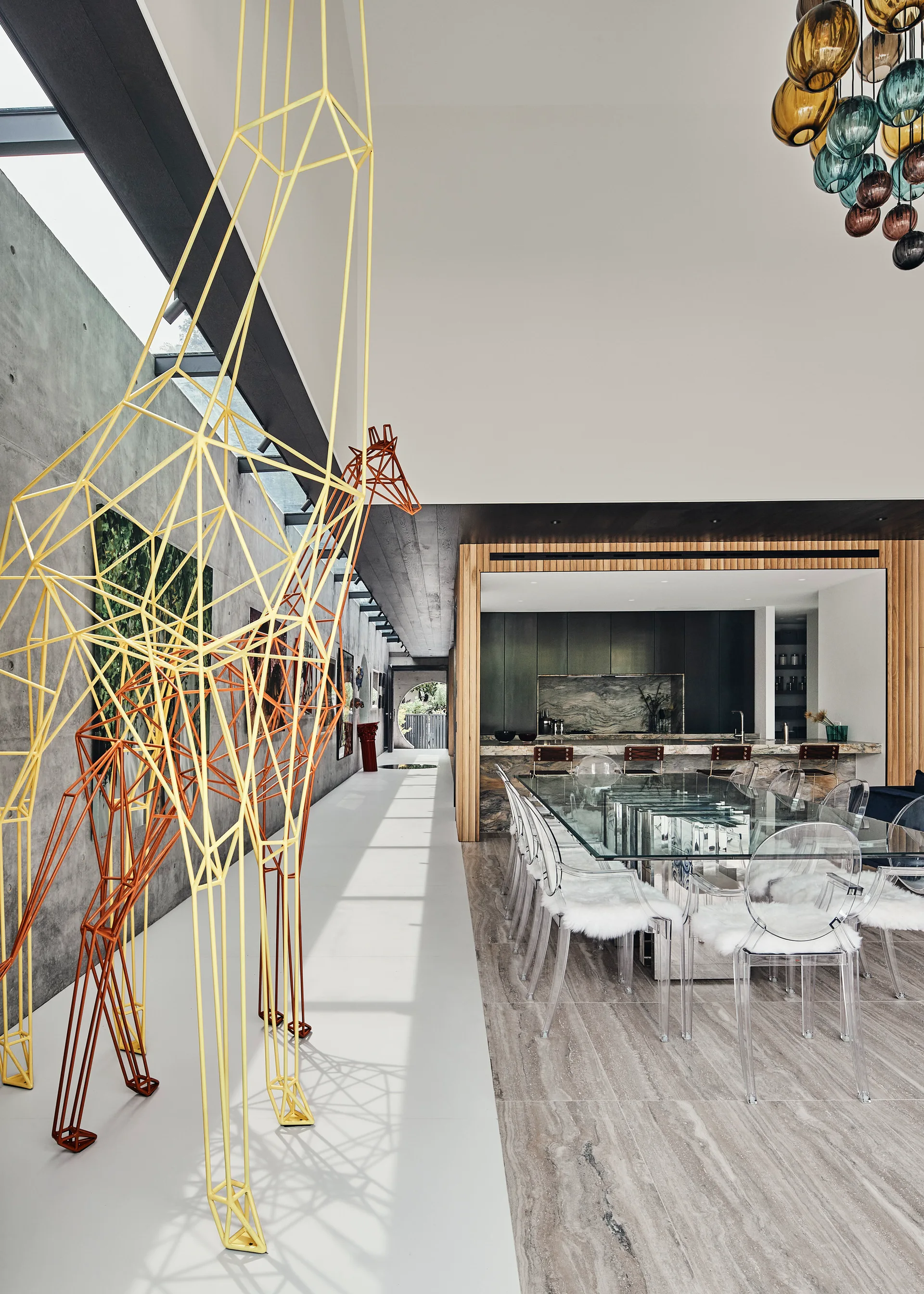 A modern dining room features a glass table with transparent chairs and large wire giraffe sculptures next to a grey concrete wall.