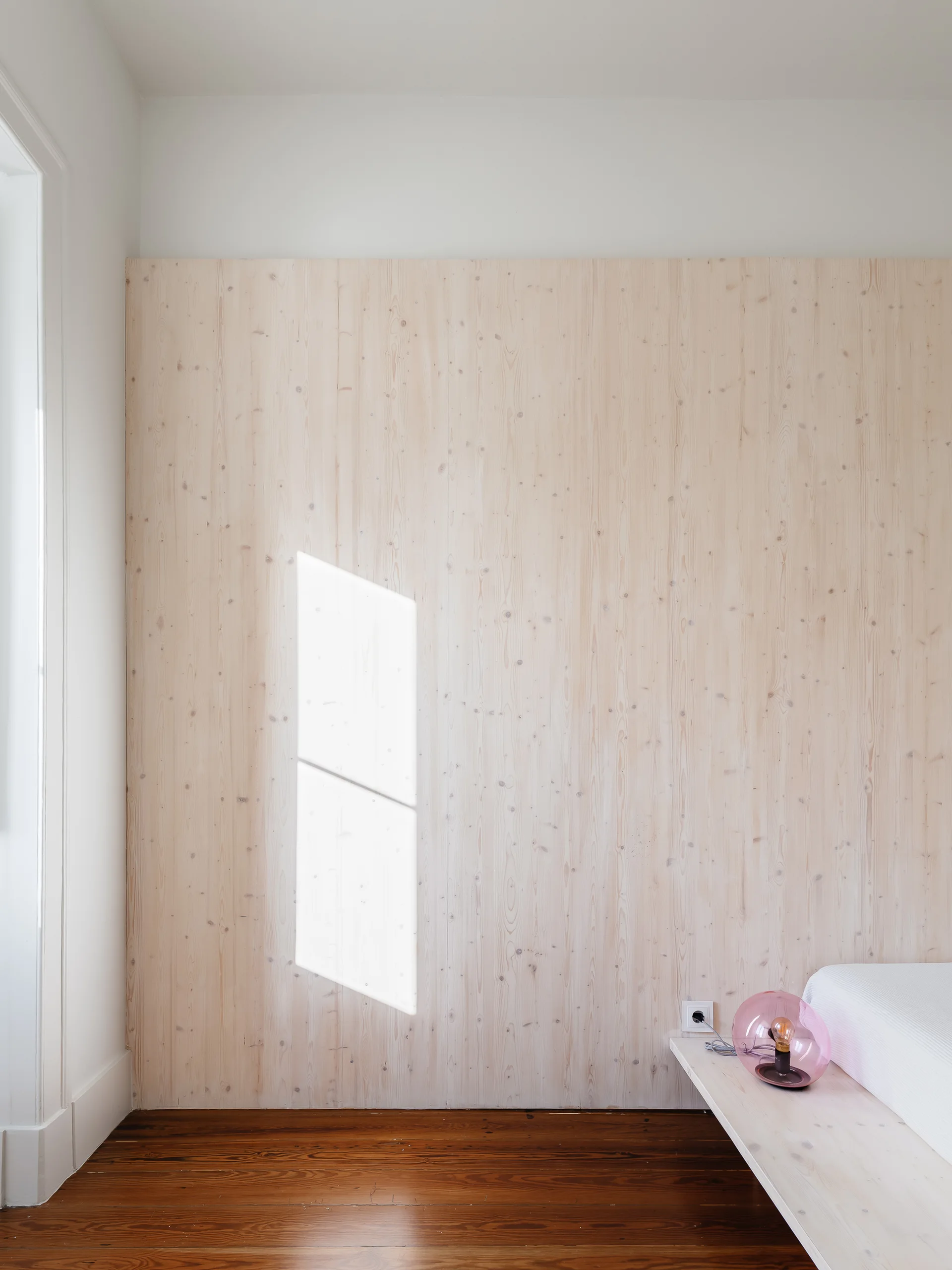 A minimalist bedroom corner featuring a light wood paneled accent wall, a low floating wood platform shelf, and a pink glass globe lamp.