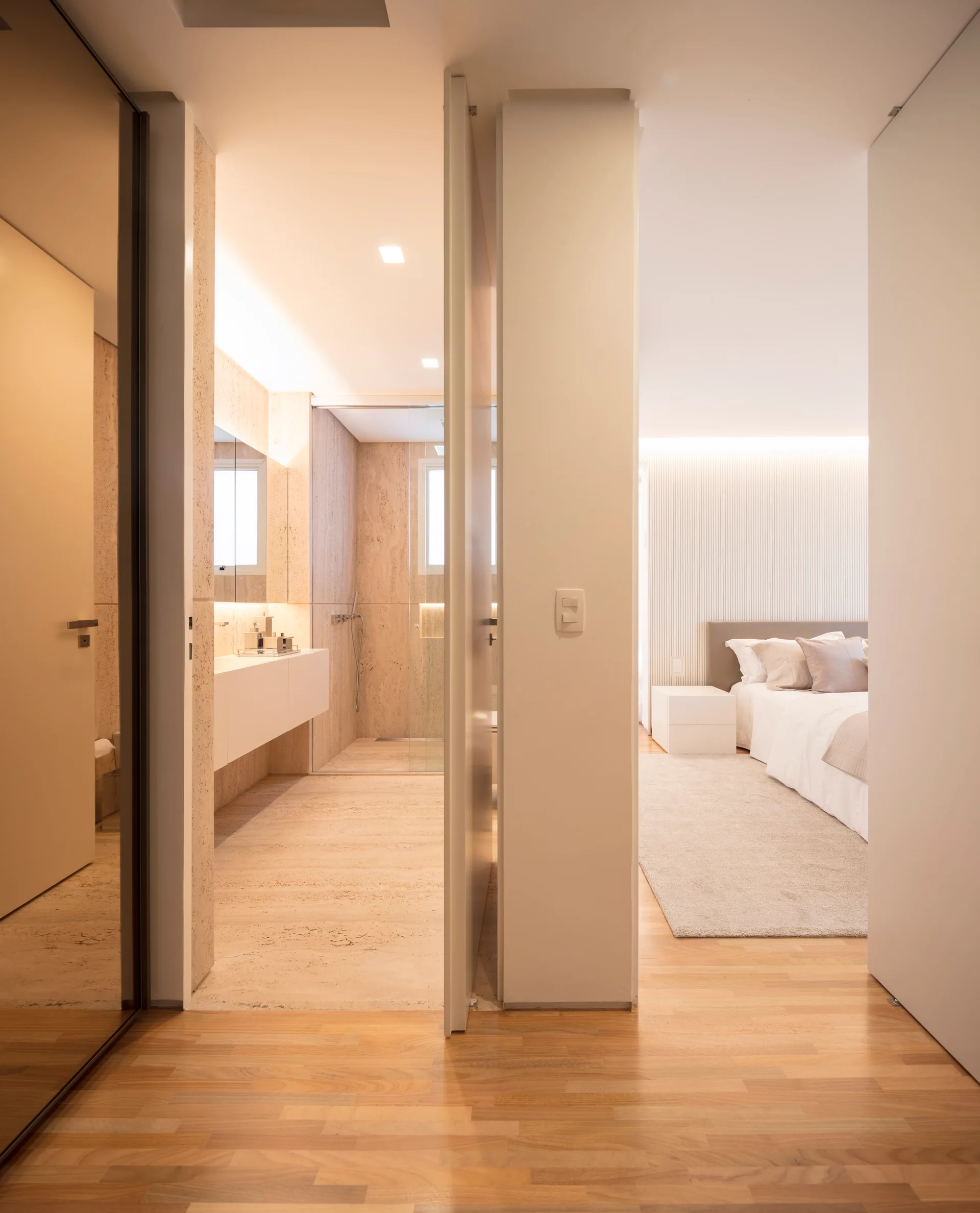 An open doorway reveals a warm-toned bedroom with a slatted accent wall adjacent to a travertine stone bathroom featuring a floating white vanity.