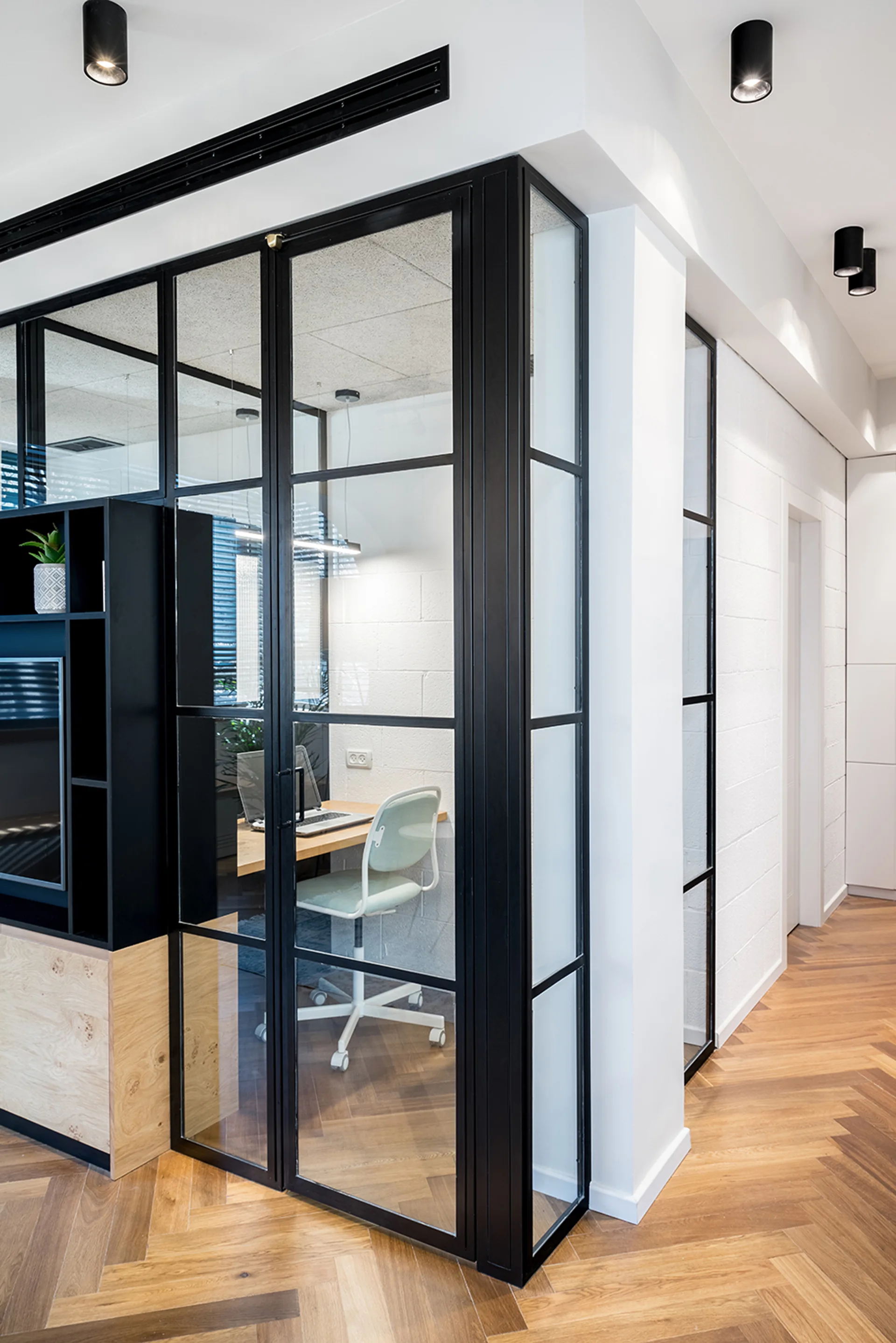 A home office enclosure with a black steel-framed glass partition, wooden desk, white swivel chair, and herringbone hardwood flooring.
