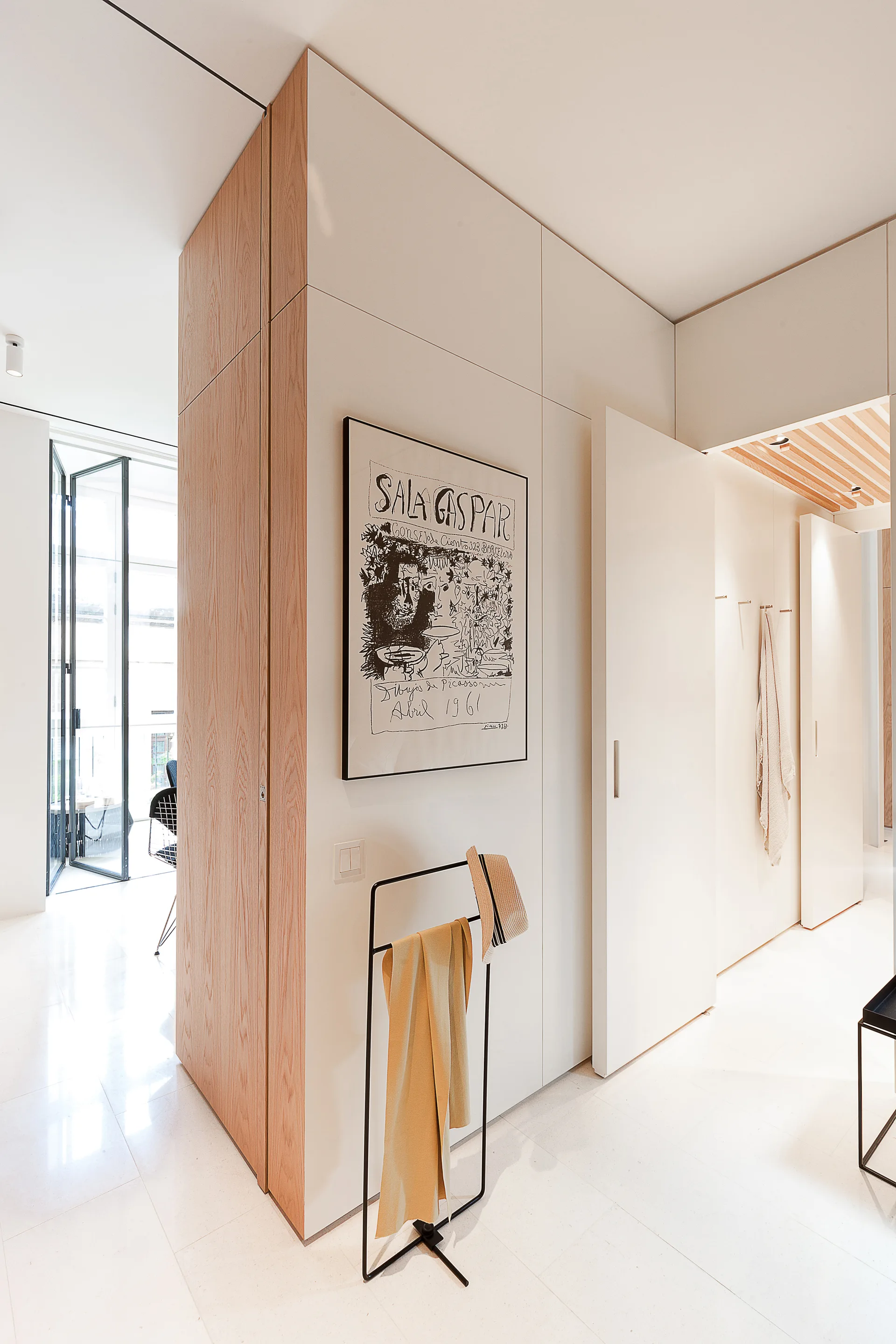 A modern entryway features white flat-front storage cabinets, a natural oak corner panel, a black metal coat rack, and polished white floors.