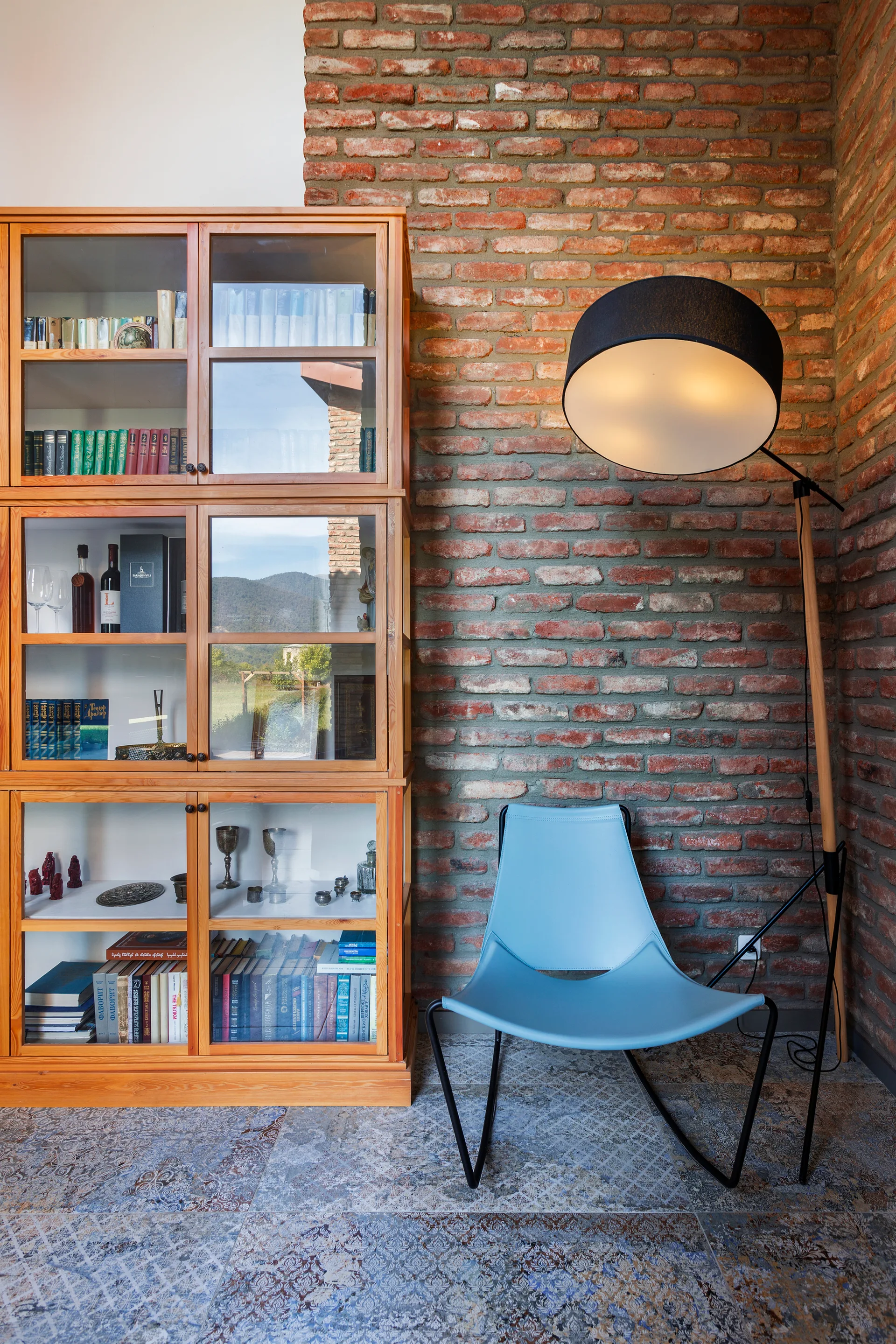 A reading corner features a wooden glass-fronted bookcase, a light blue modern chair, and an arched floor lamp against an exposed brick wall.