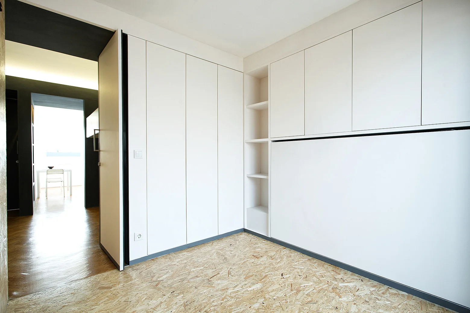 An empty room features handleless white built-in cabinets, a narrow column of open shelving, and textured OSB flooring.