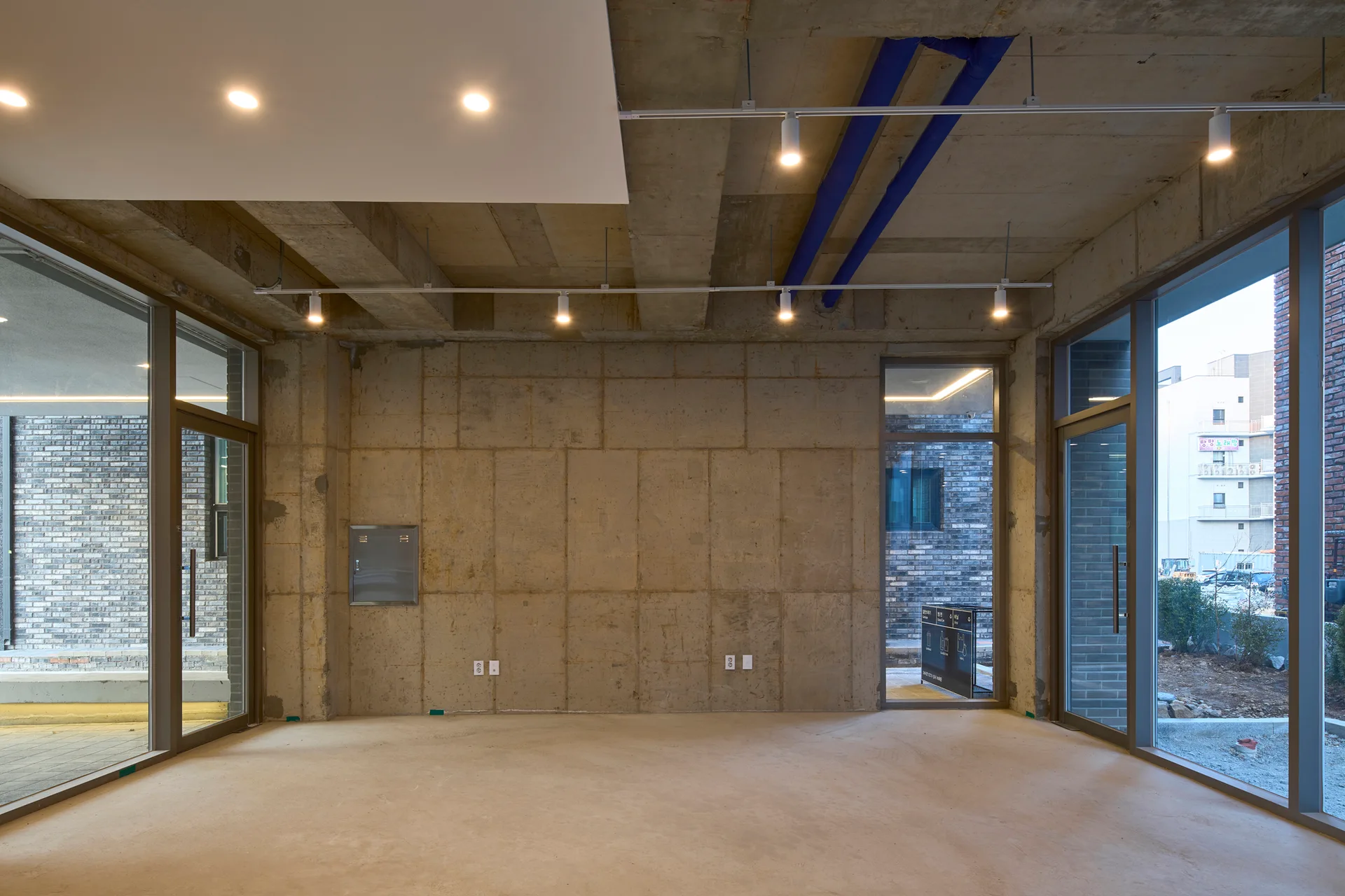 An empty industrial space features bare concrete walls and ceilings, track lighting, exposed blue pipes, and large metal-framed windows.