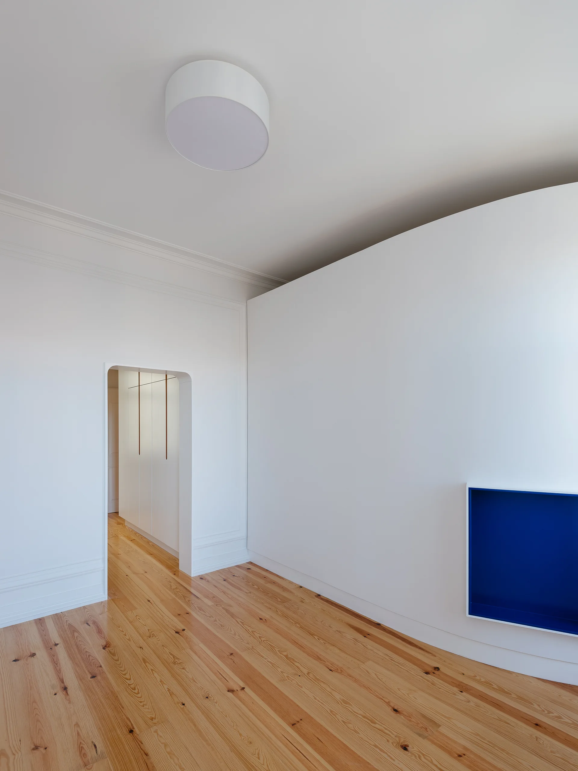 A minimalist white room features a vibrant blue recessed wall niche, a curved doorway leading to a hallway, and warm pine hardwood floors.