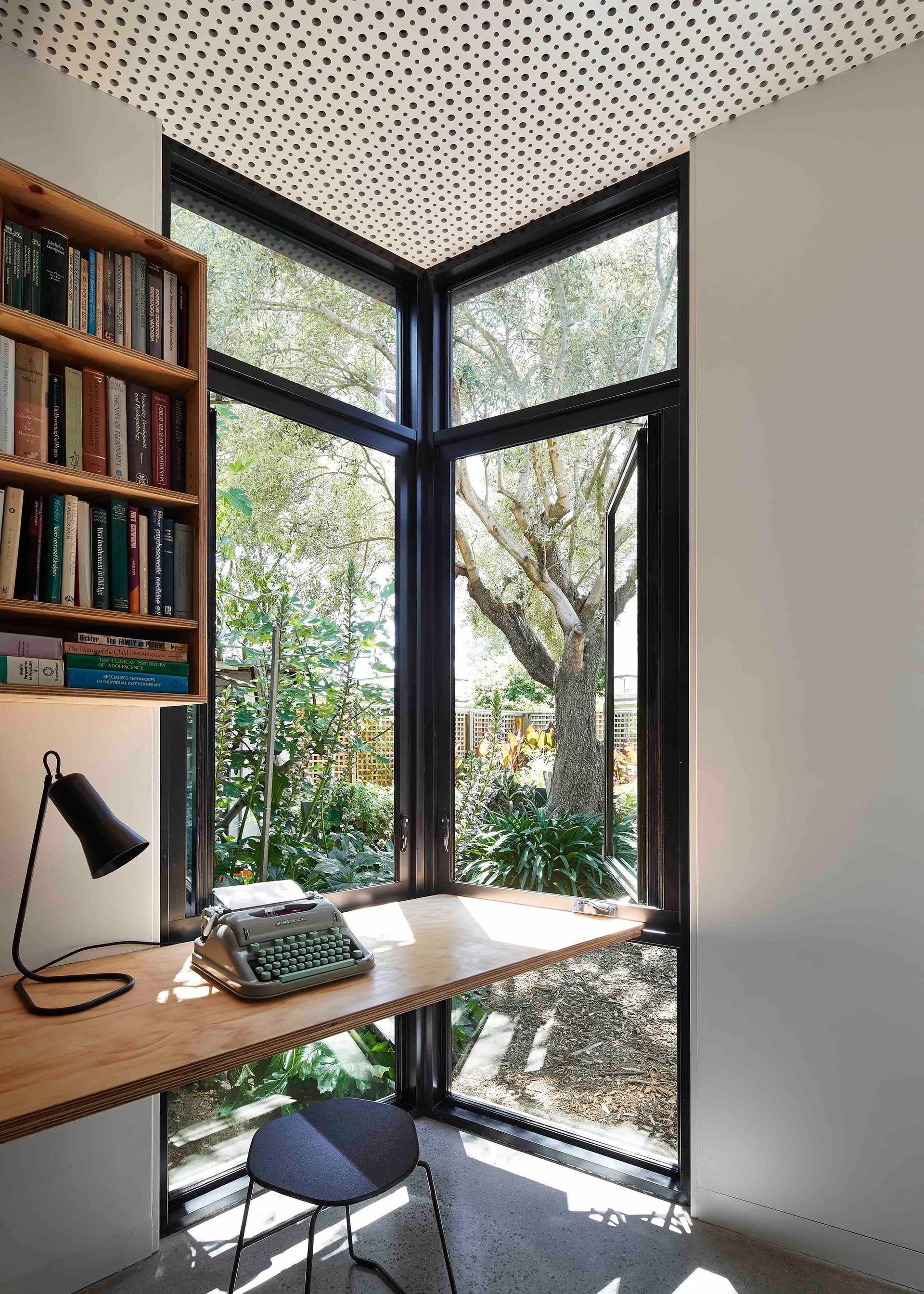 A minimalist home office features a plywood desk and built-in bookshelf set against a large black-framed corner window overlooking a garden.