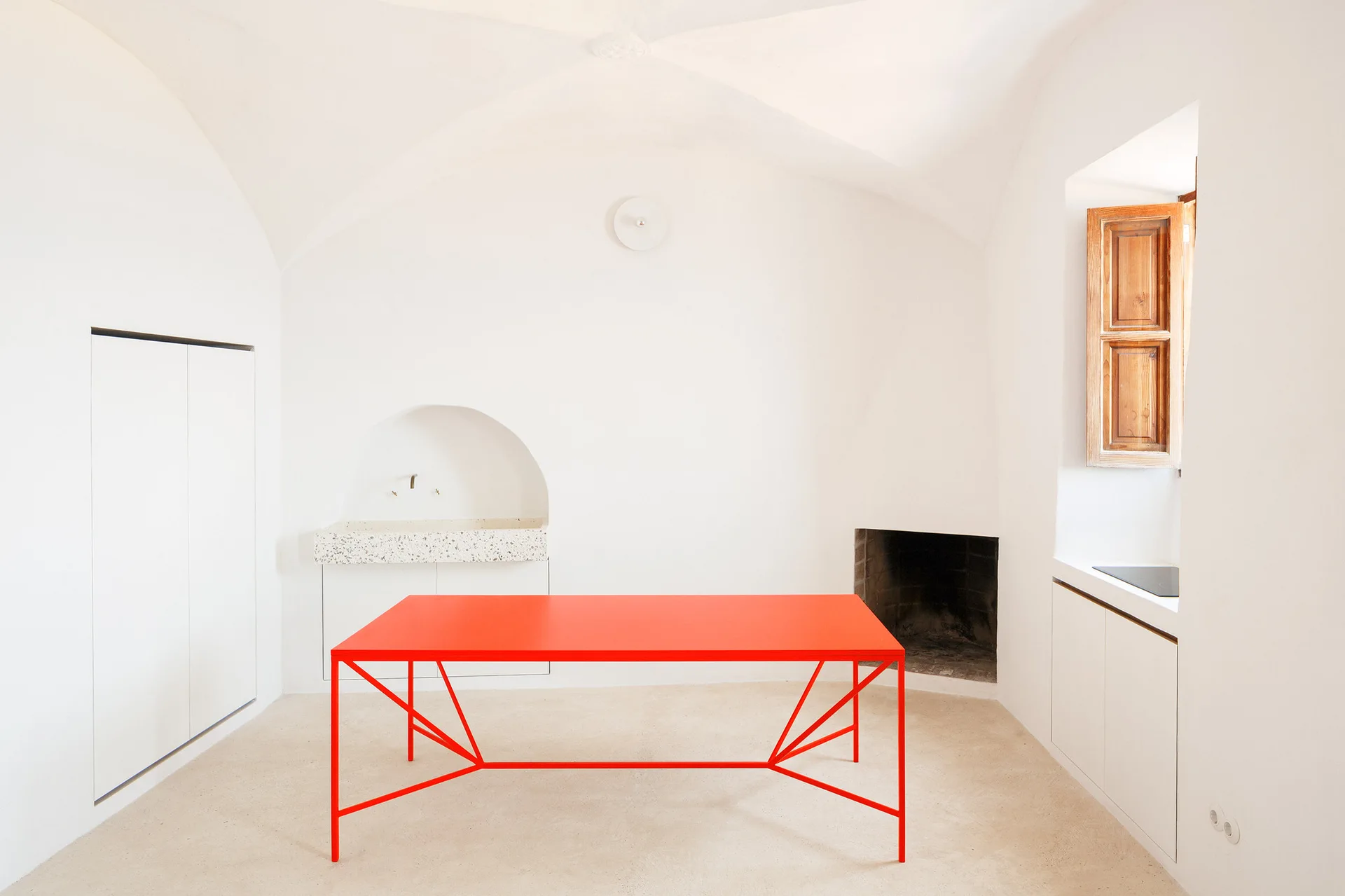 A minimalist white kitchen with a vaulted plaster ceiling, an arched alcove sink, wooden shutters, and a bright red rectangular table.