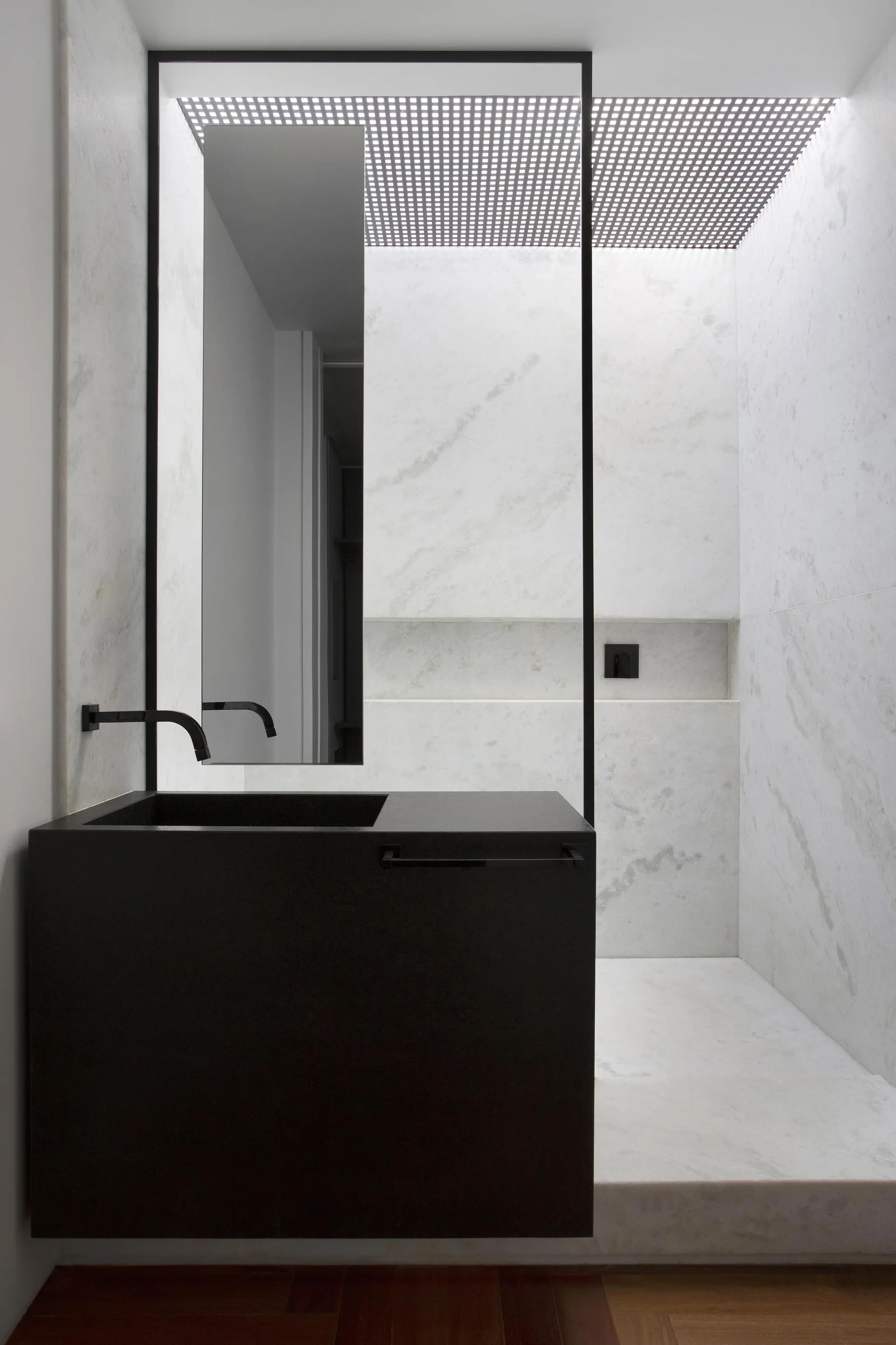 A modern bathroom features a matte black block vanity and faucet against white veined marble walls with a vertical mirror and a gridded ceiling light.