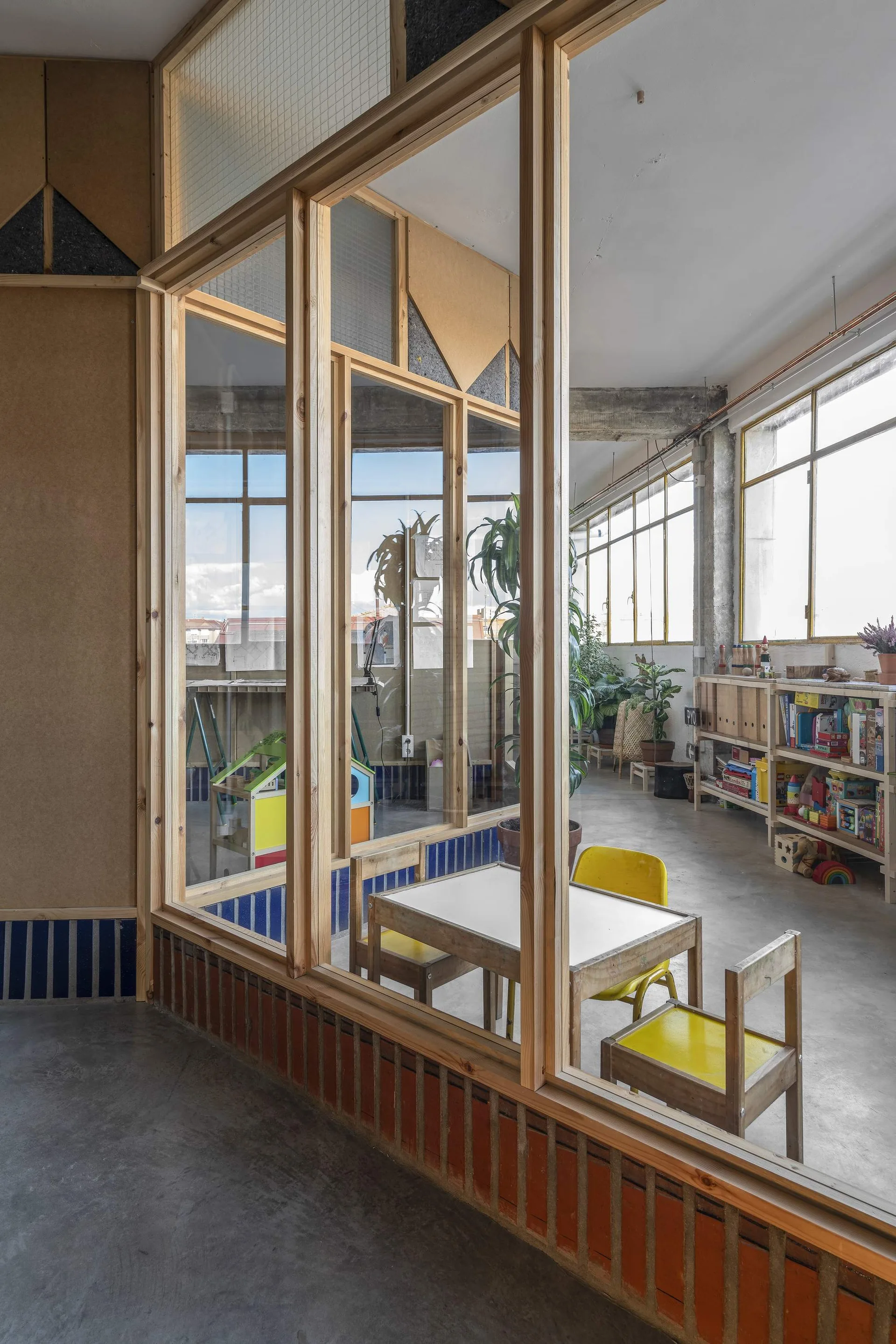 A minimalist children's playroom with a small wooden table, yellow chairs, open wood shelving, and a large timber-framed glass partition.