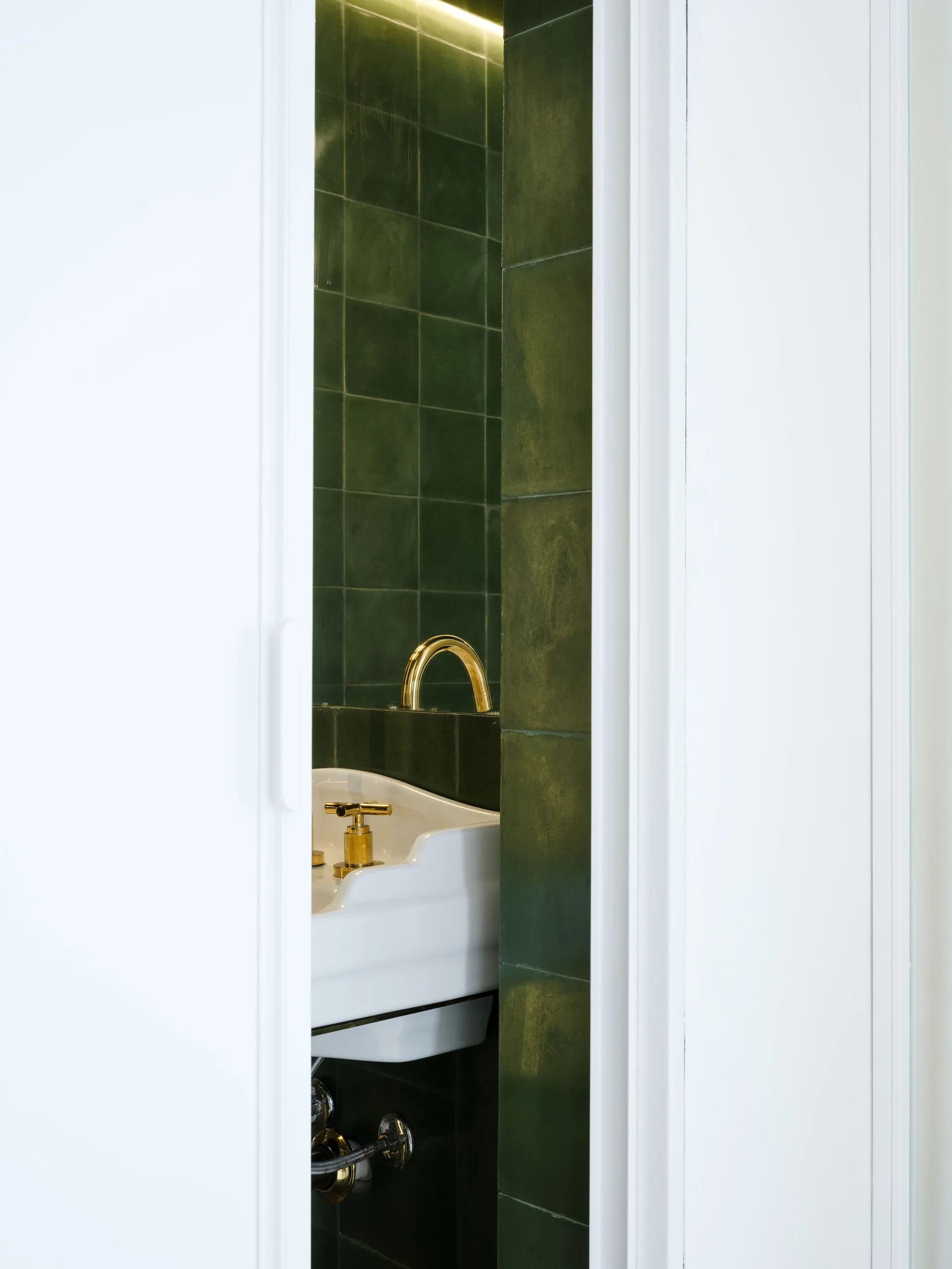 A view through a narrow white doorway reveals a bathroom sink with a gold faucet set against a dark green square-tiled wall.