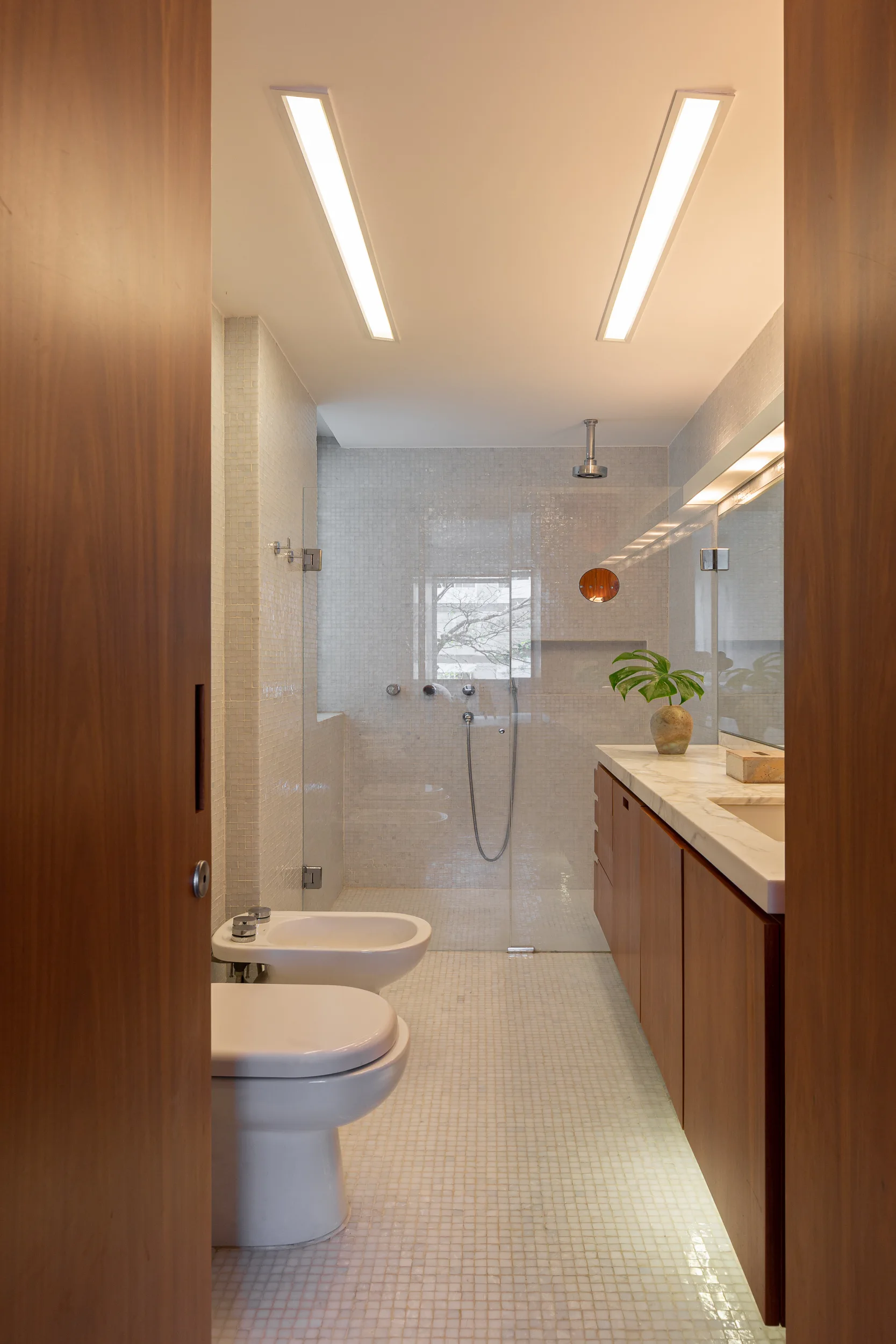 A modern bathroom features a floating wood vanity with marble countertop, a glass walk-in shower, and walls lined with small white mosaic tiles.