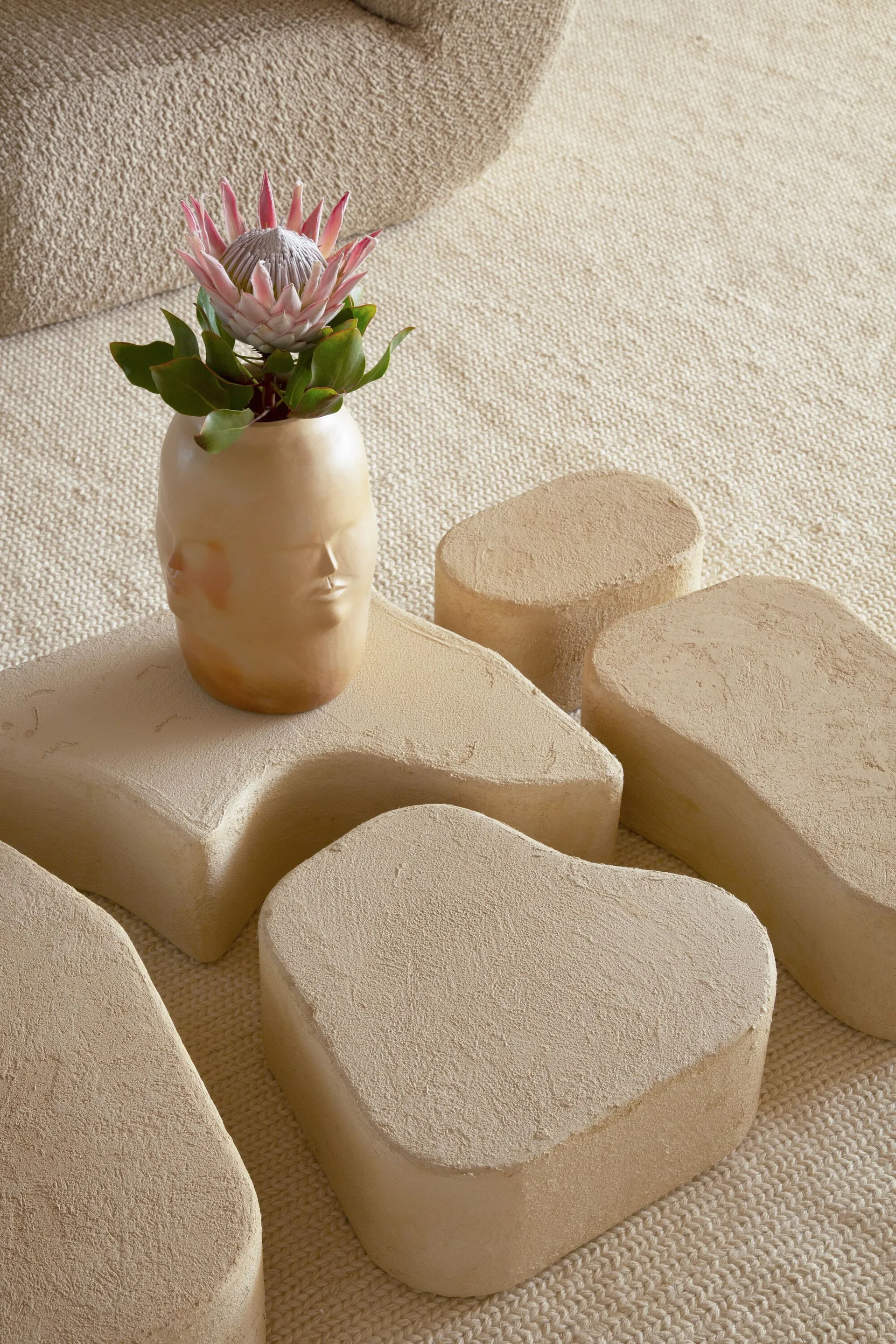 A cluster of modular beige stone blocks forming a coffee table on a woven rug, topped with a face-shaped ceramic vase containing a pink protea.