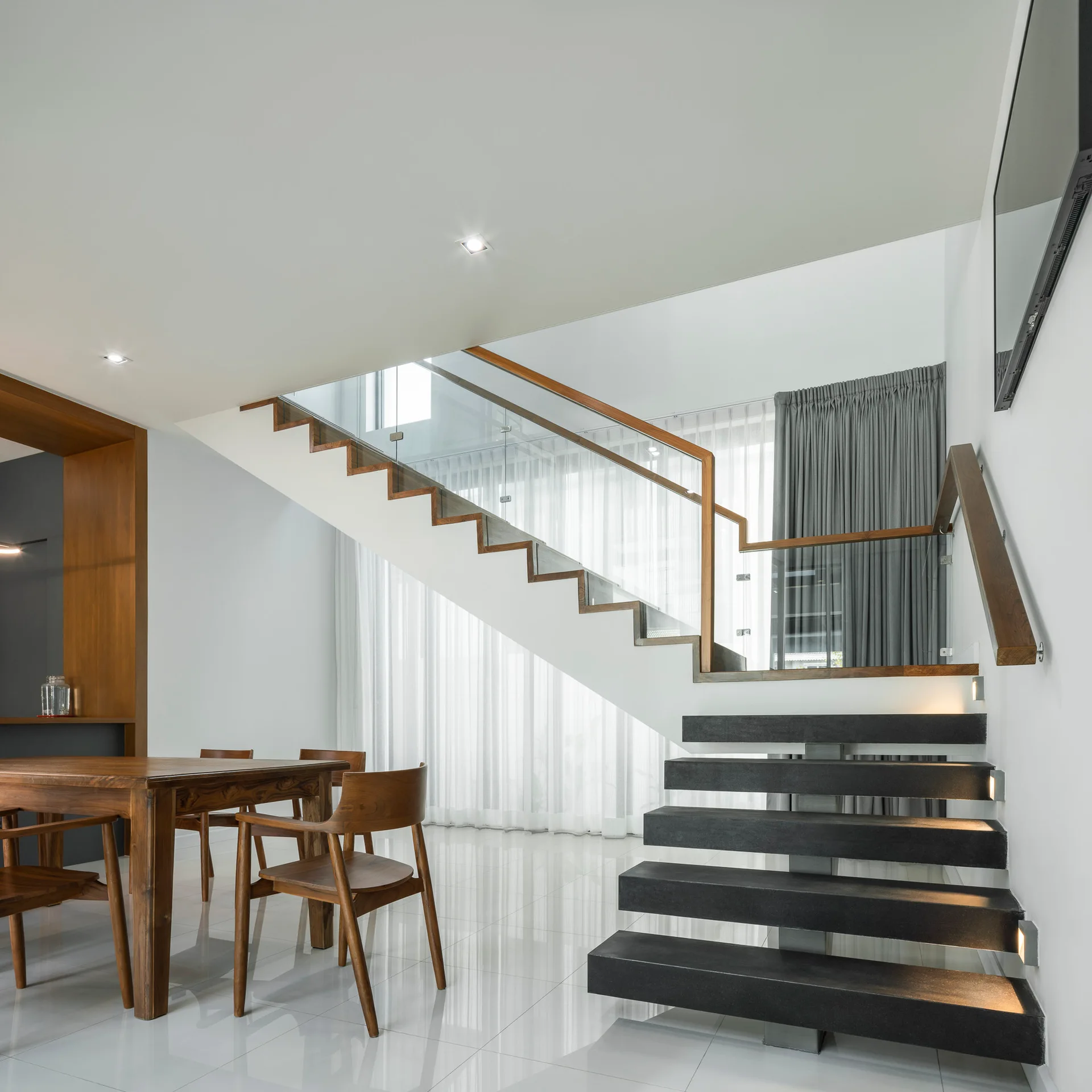 A modern white room features a floating black staircase with glass railings next to a wooden dining table set on polished white tiles.