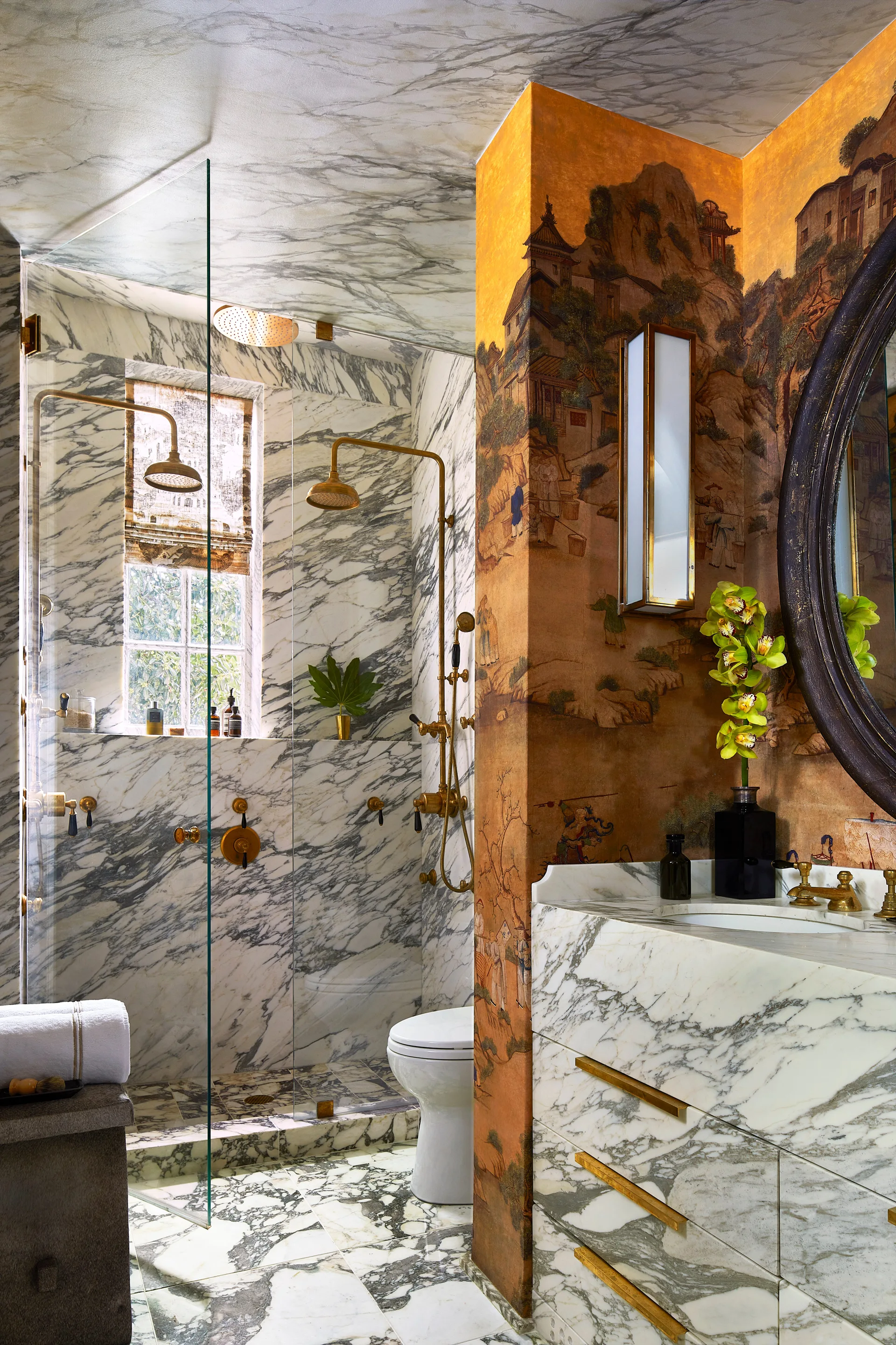 A bathroom features veined white marble on the shower walls, floor, and vanity drawers, paired with scenic gold wallpaper and brass hardware.