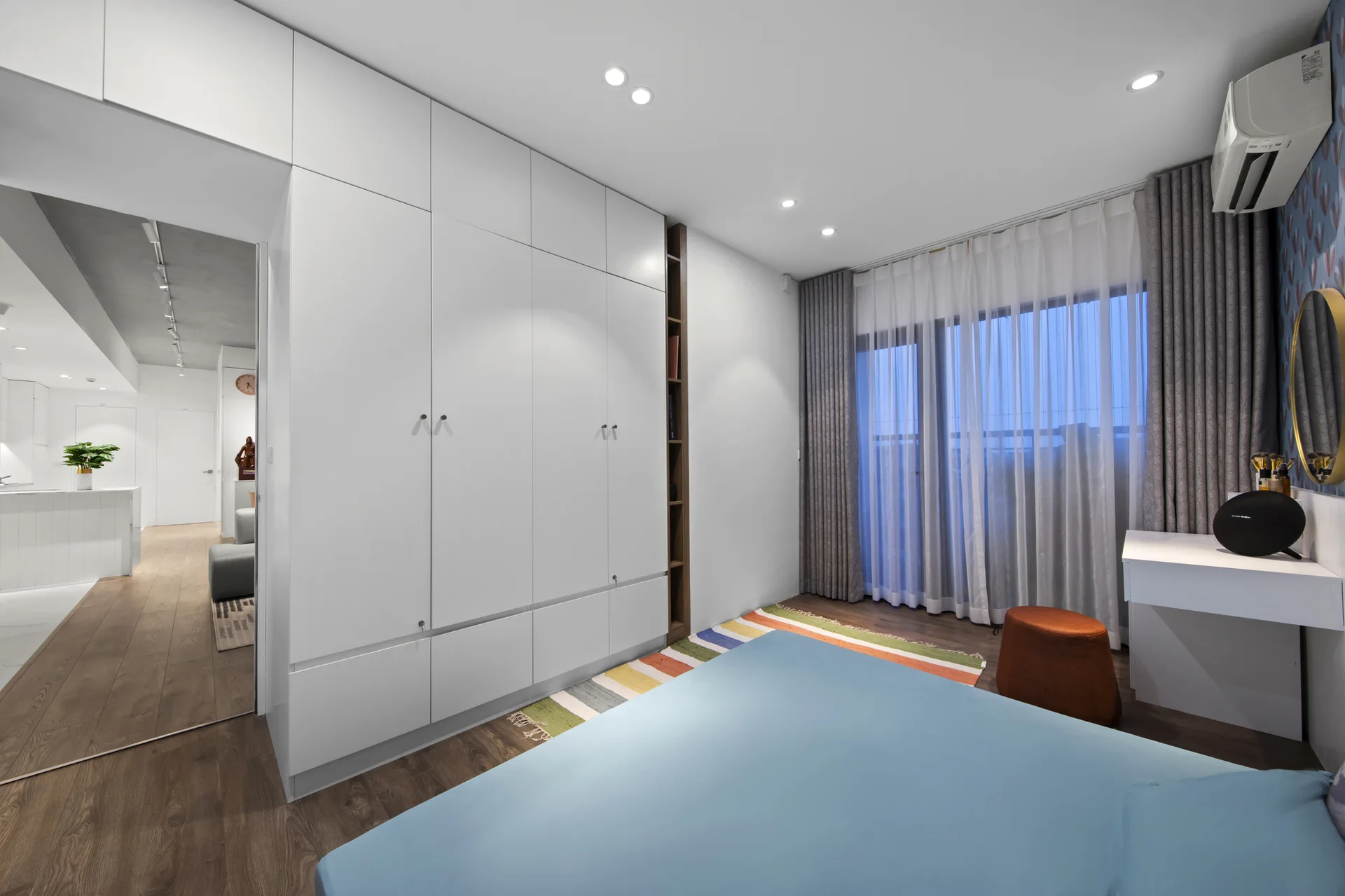 A modern bedroom features a large white floor-to-ceiling wardrobe, a bed with blue linens, a colorful striped rug, and a small white vanity.