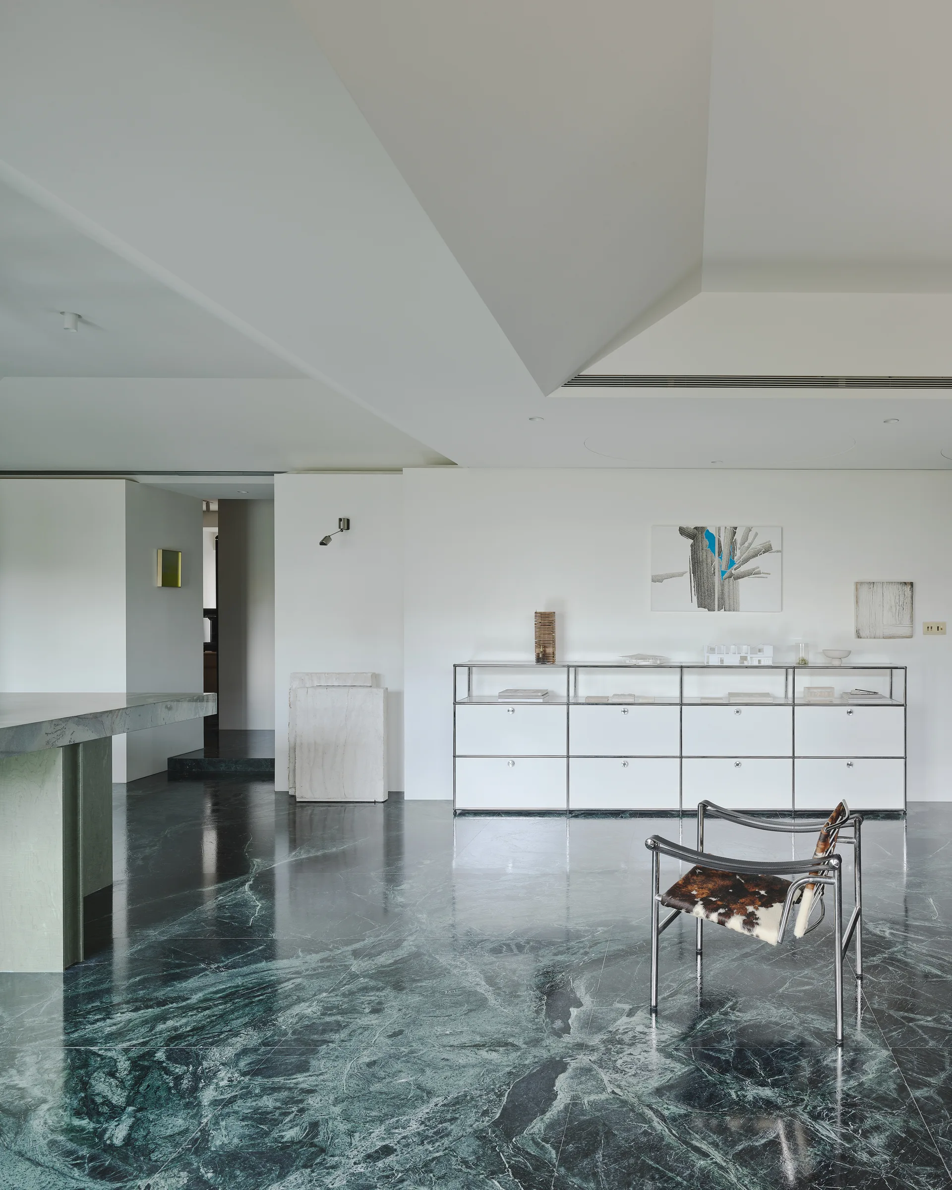 A minimalist room features a polished dark green marble floor, a white modular cabinet against the wall, and a chrome-framed hide chair.