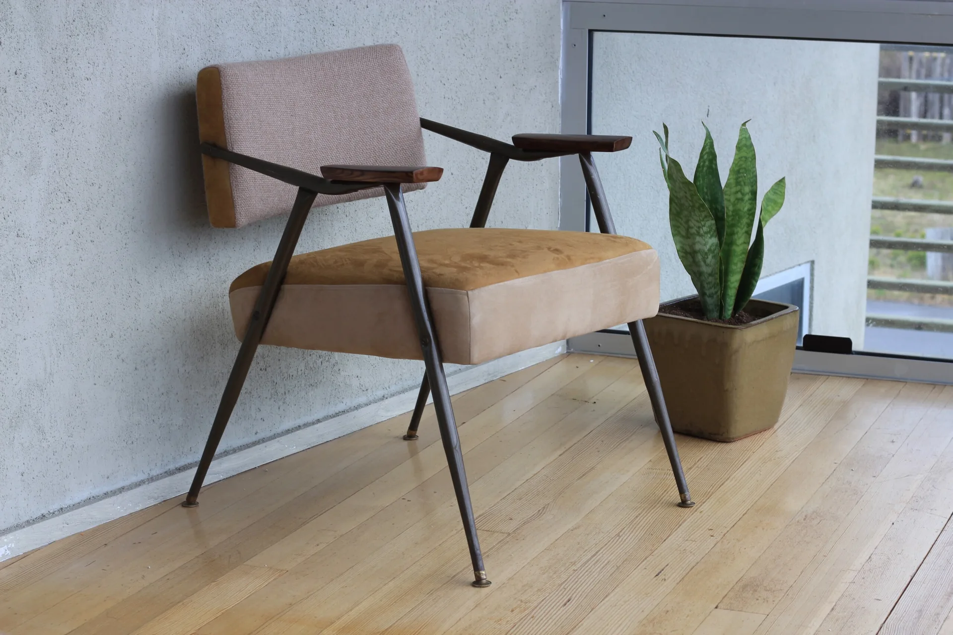 A mid-century modern armchair with two-tone upholstery stands on a light wood floor next to a potted snake plant.