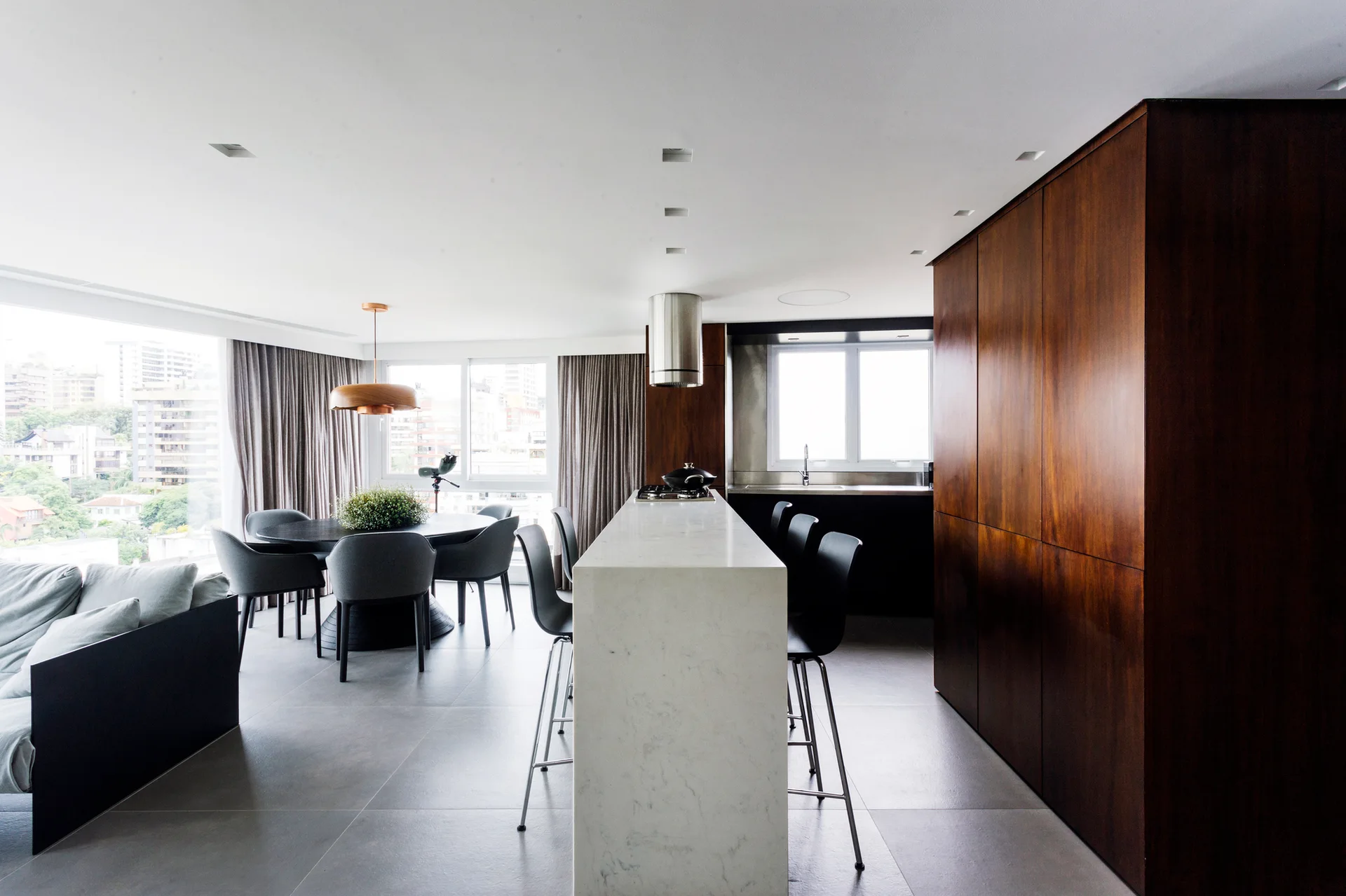 A modern kitchen featuring a white marble island with stools, a floor-to-ceiling dark wood cabinet wall, and an adjacent dining area.