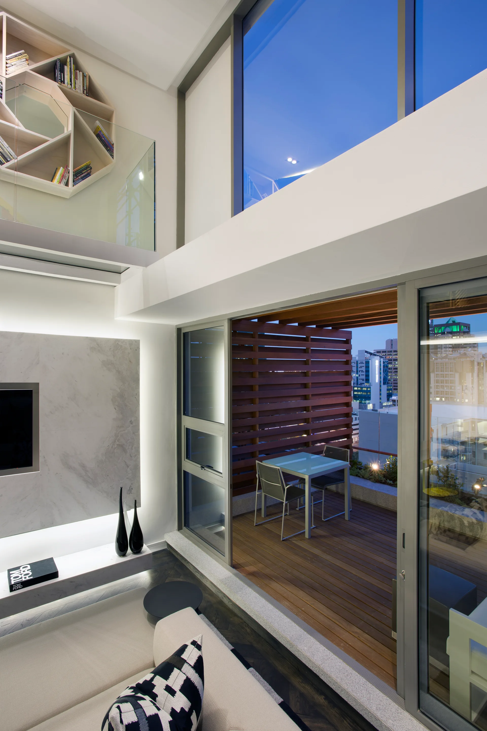 A modern double-height living room opens to a wood-decked balcony with a small dining table, featuring a mezzanine level with a geometric bookshelf.