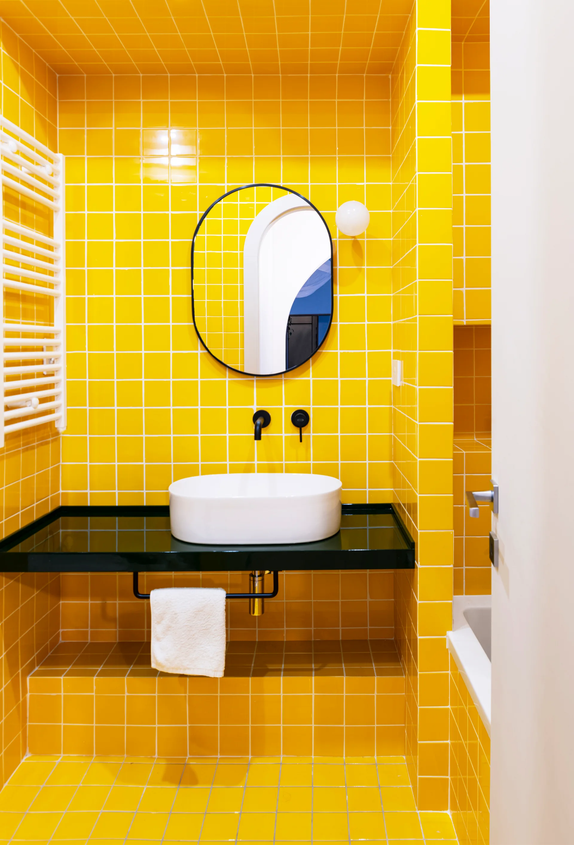 A modern bathroom featuring bright yellow grid-patterned tile walls and floor, a black floating vanity with a white vessel sink, and an oval mirror.