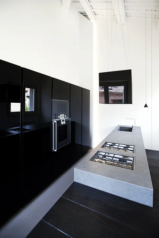 A minimalist kitchen featuring reflective black cabinetry, a matte grey stone island with a built-in cooktop, and painted white ceiling beams.