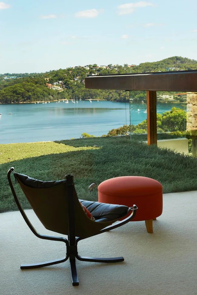 A modern dark-frame armchair and a round orange ottoman sit on off-white carpet in front of a floor-to-ceiling glass wall overlooking a bay.
