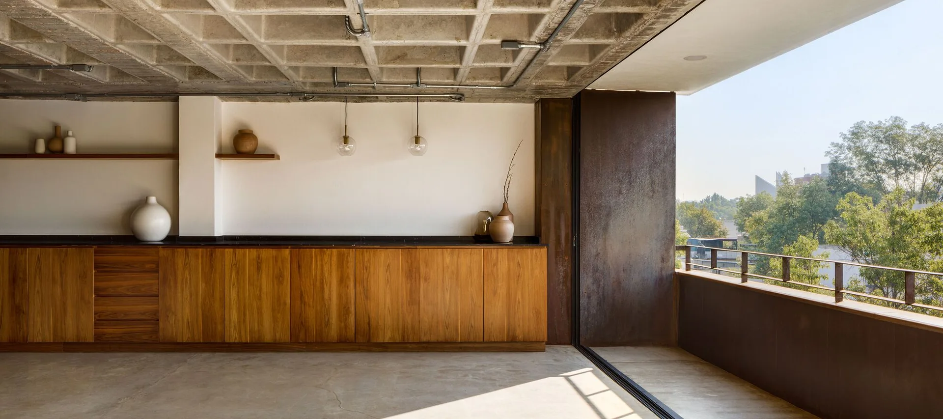 A long wooden kitchen cabinet with a black countertop stands beneath an exposed concrete coffered ceiling next to a sunny balcony.