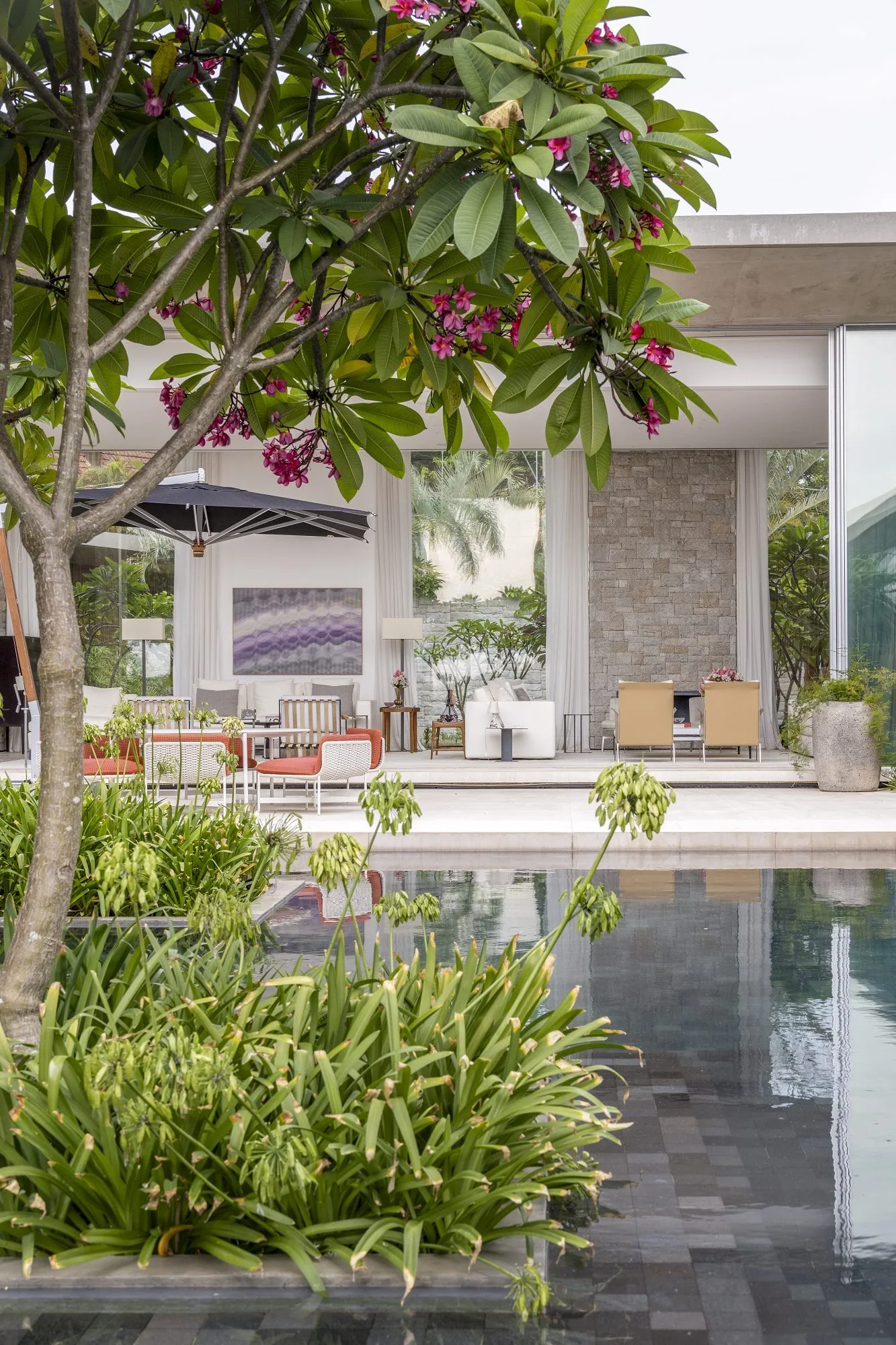 A contemporary poolside pavilion features white outdoor seating, a stacked stone wall, and a foreground of lush green foliage and a flowering tree.