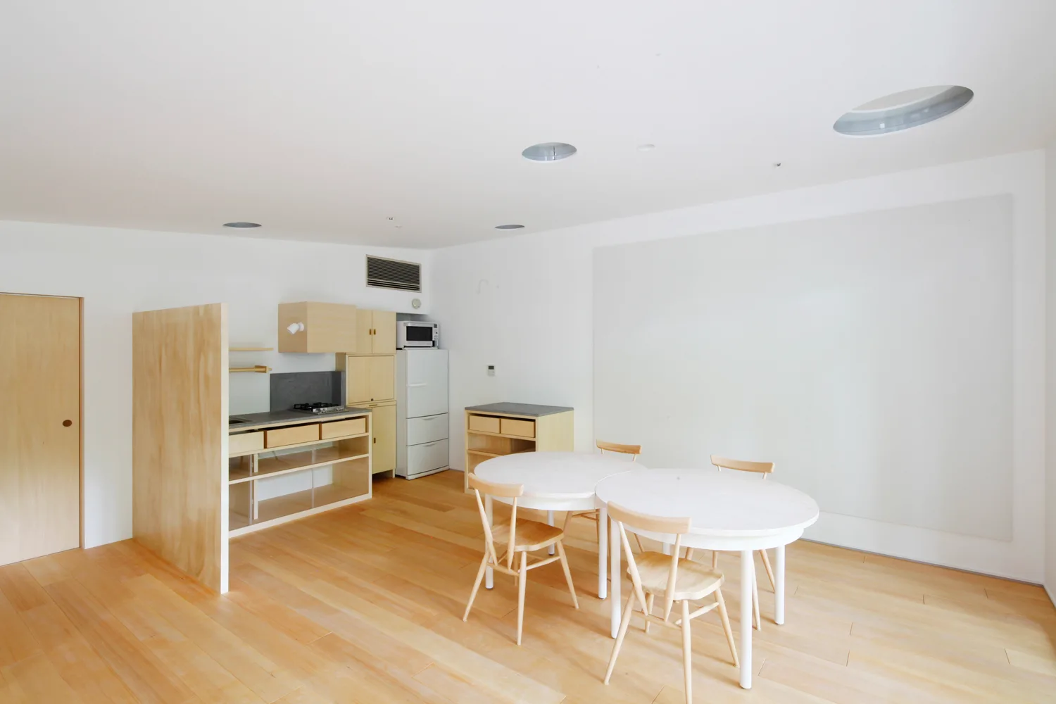 A minimalist white kitchen and dining space featuring a light wood partition wall, two white round tables, and light wood flooring.