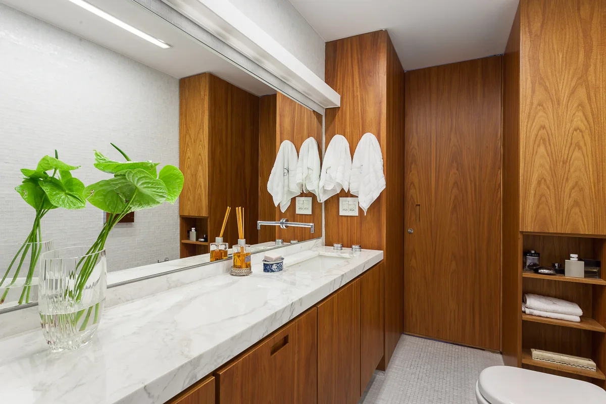 A modern bathroom vanity features a white veined marble countertop, warm wood cabinetry, a large mirror, and white mosaic tiled walls.
