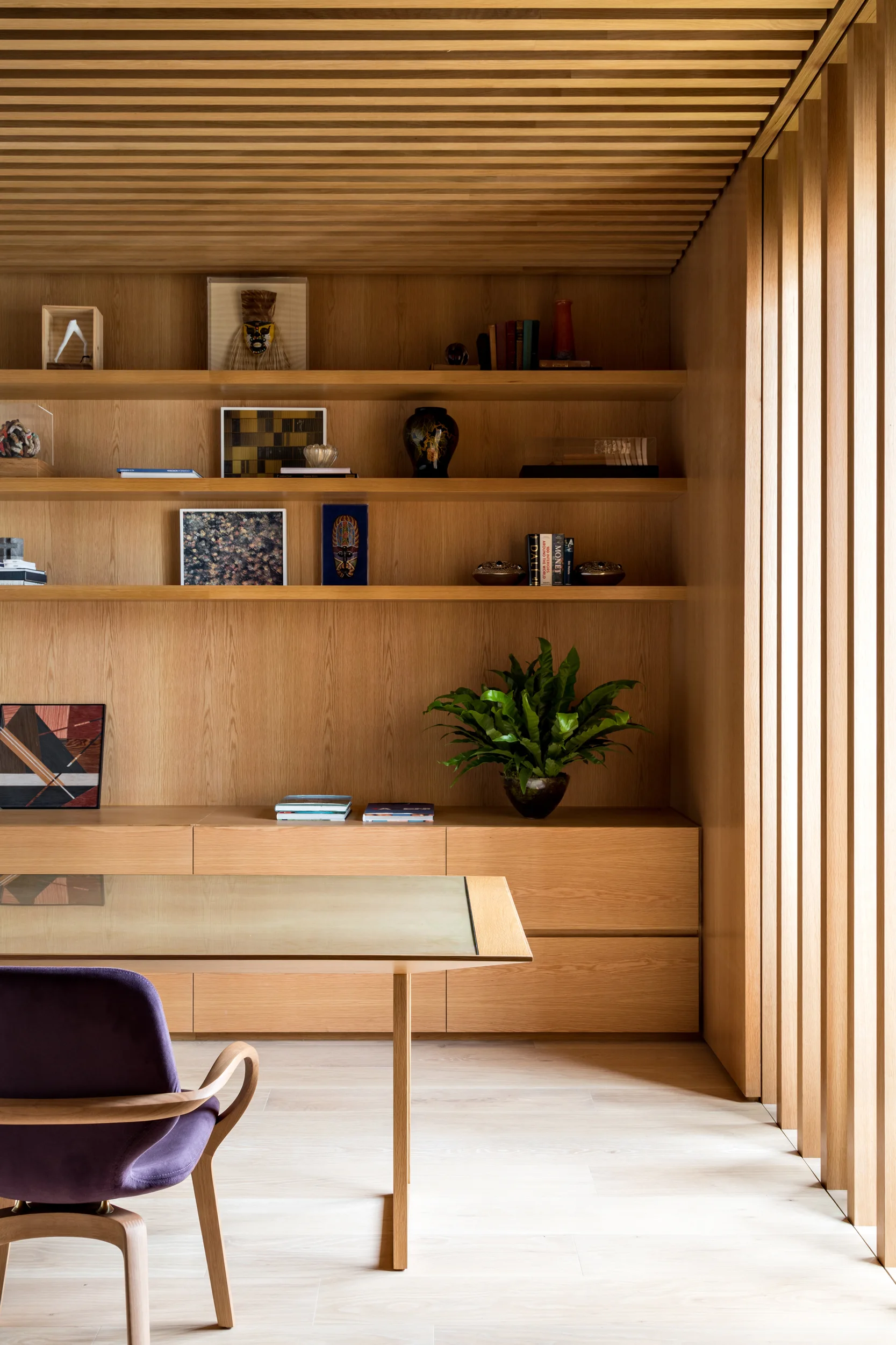 A home office features light wood-paneled walls, a matching slatted ceiling, built-in shelves with books and decor, and a purple upholstered desk chair.