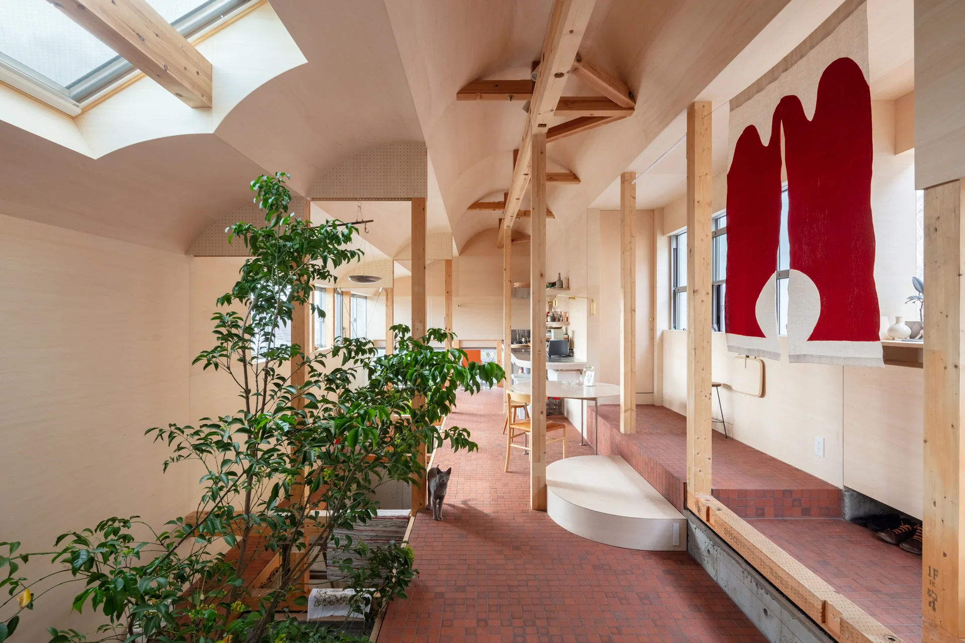 An open residential interior features a vaulted wood ceiling, exposed timber posts, a tiered terracotta tile floor, and a red wall hanging.