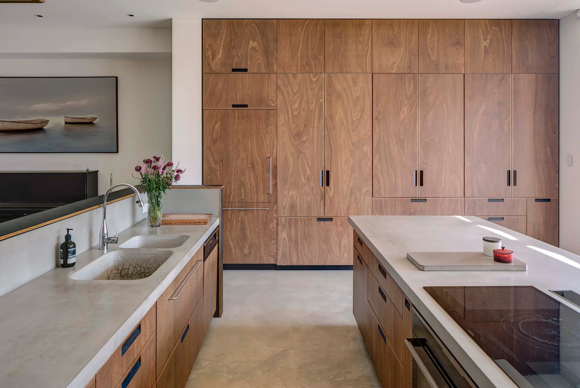 A modern kitchen featuring a wall of rich wood veneer cabinets, a central island, concrete countertops, and an integrated double sink.