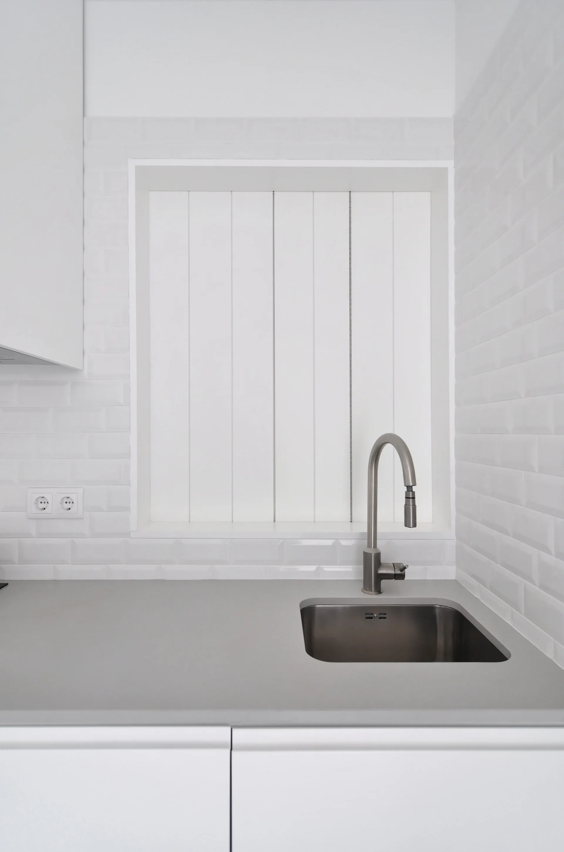 A stainless steel kitchen sink with a gooseneck faucet is set in a grey countertop against a white subway tile backsplash and vertical window shutters.