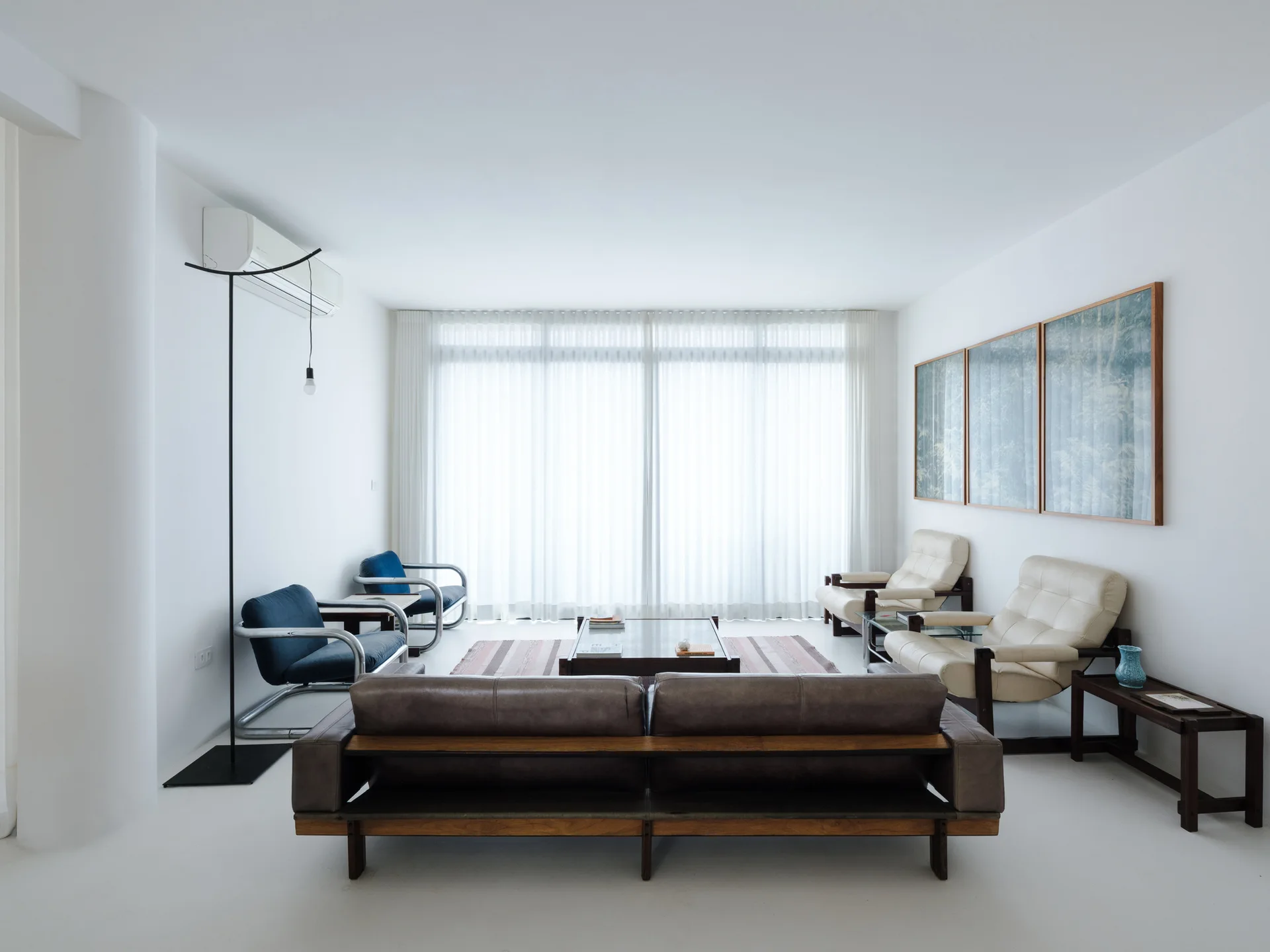 A minimalist white living room features a dark leather sofa, blue and cream armchairs, a low wooden coffee table, and a large window with sheer curtains.