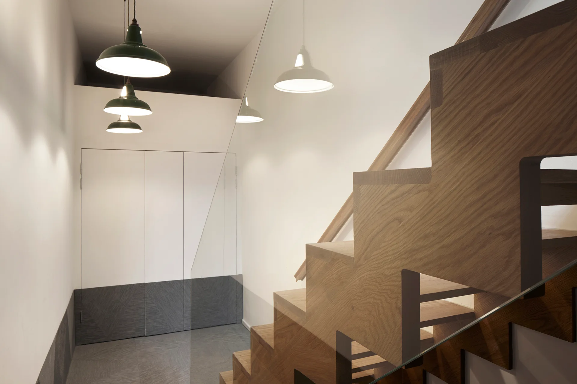 A modern wooden staircase with a transparent glass railing is illuminated by three dark green pendant lights next to a white and gray built-in cabinet.
