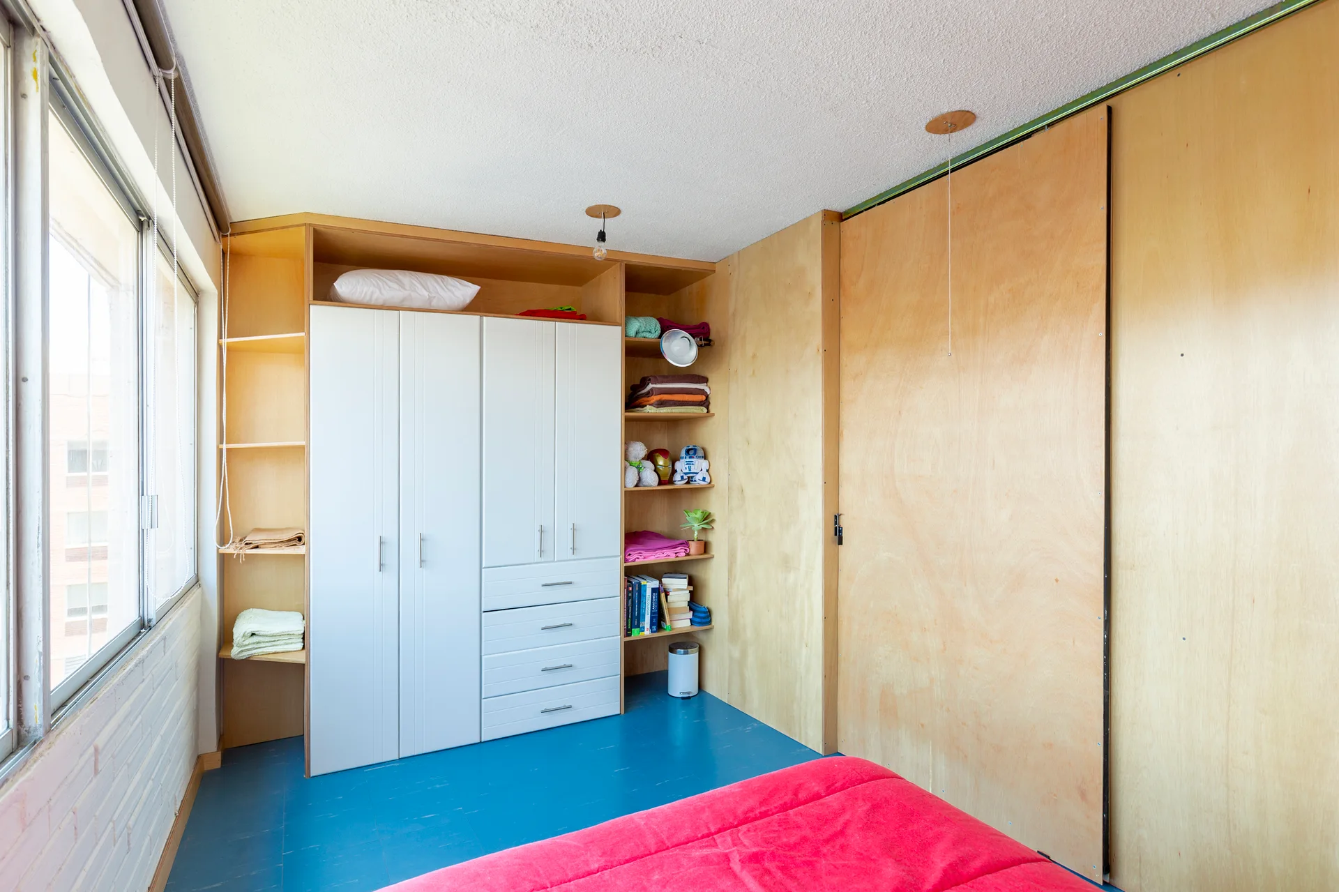 A minimalist bedroom featuring a built-in plywood wardrobe with white cabinet doors, a painted blue floor, and a red bed.