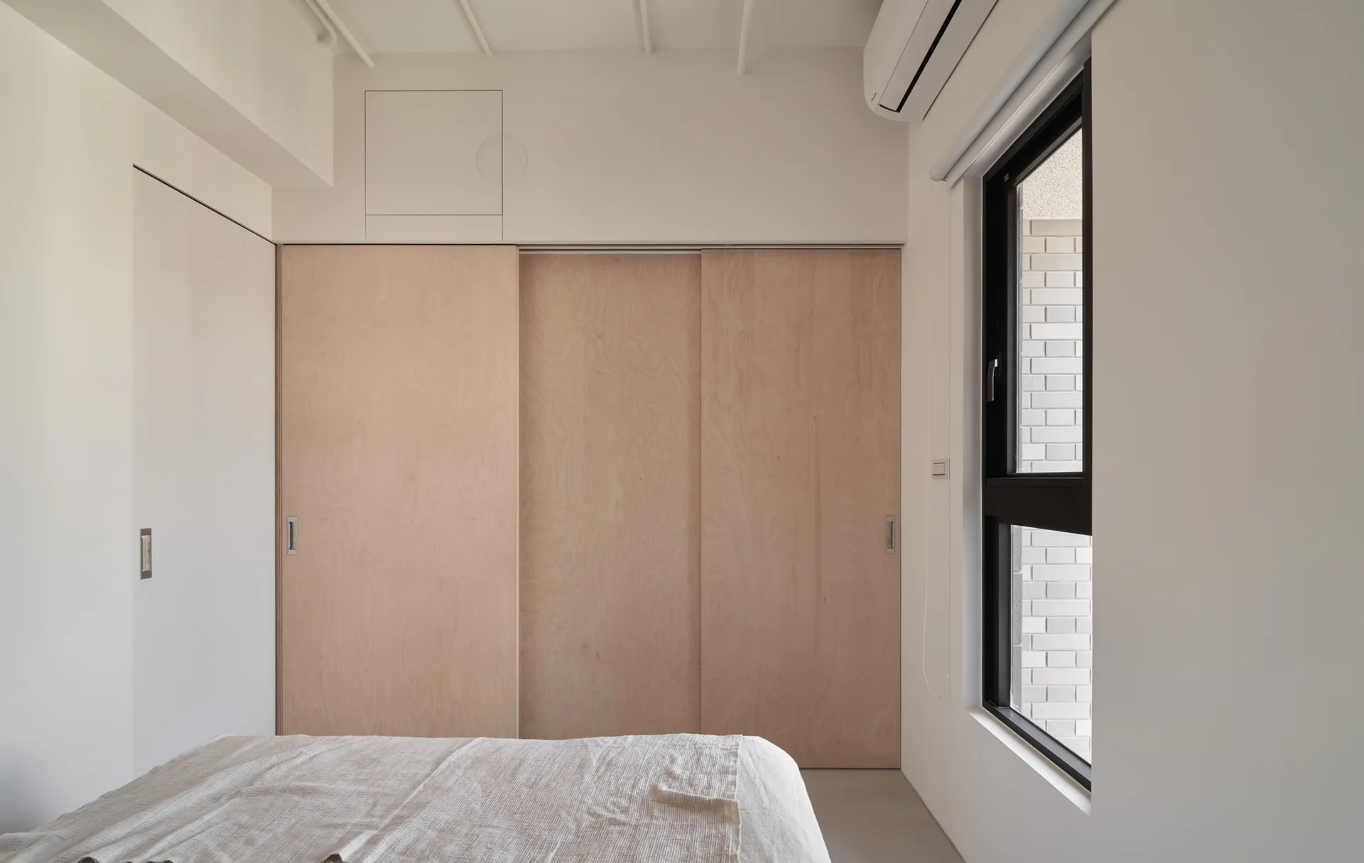 A minimalist bedroom features a bed in the foreground, large light wood sliding closet doors, and a black-framed window on a plain white wall.
