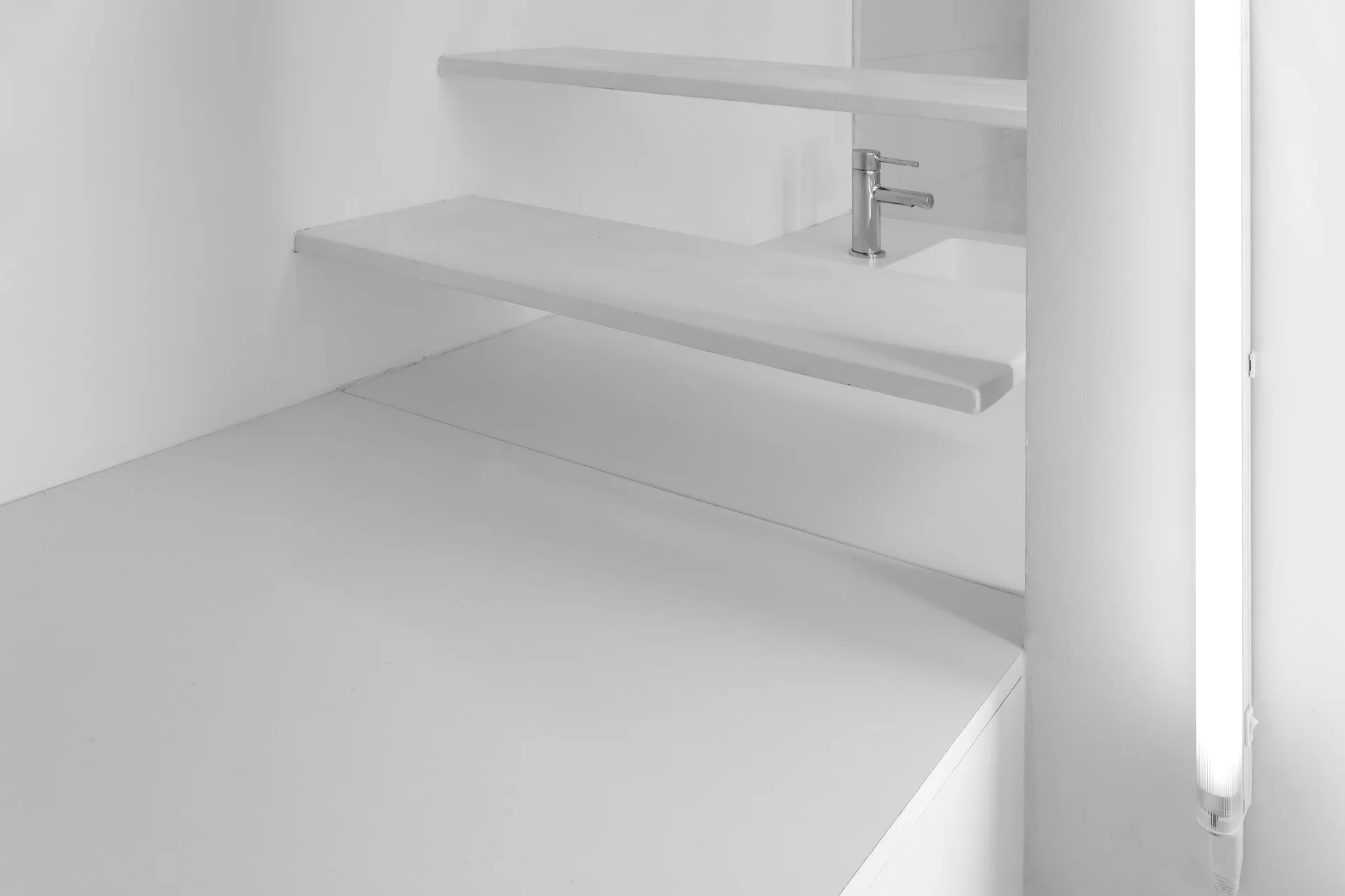 A minimalist white bathroom corner features two floating shelves, an integrated washbasin with a chrome faucet, and a vertical tube light.