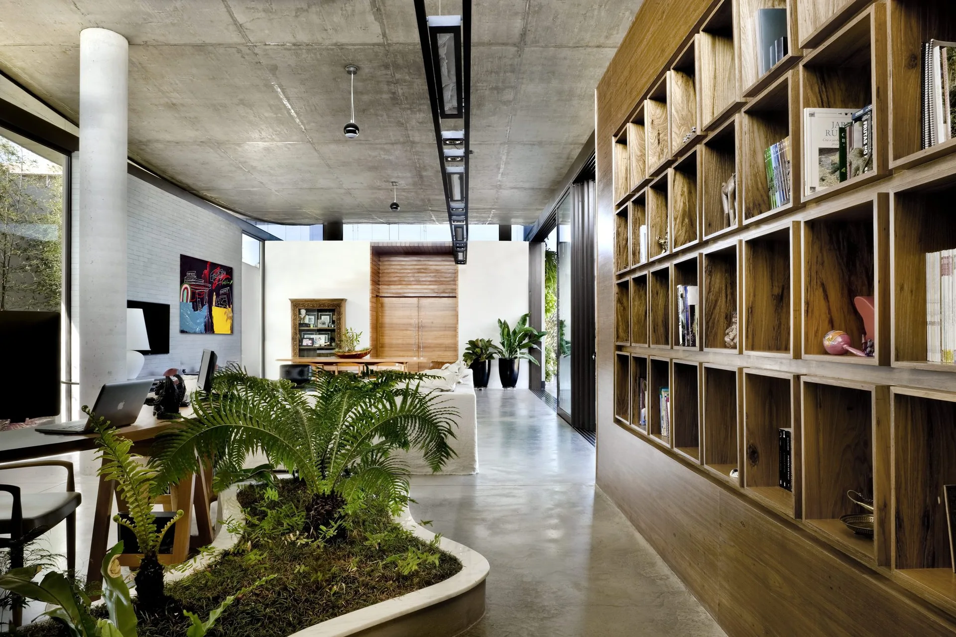 A spacious living room features an exposed concrete ceiling, a large wood grid bookshelf, a polished concrete floor, and an integrated indoor planter.