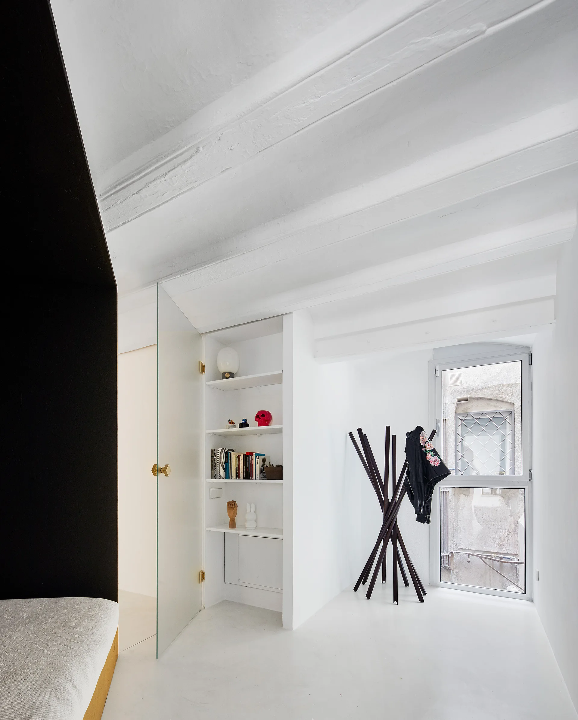A minimalist white entryway featuring painted ceiling beams, a recessed bookshelf, a glass door, and an angular black coat rack next to a window.