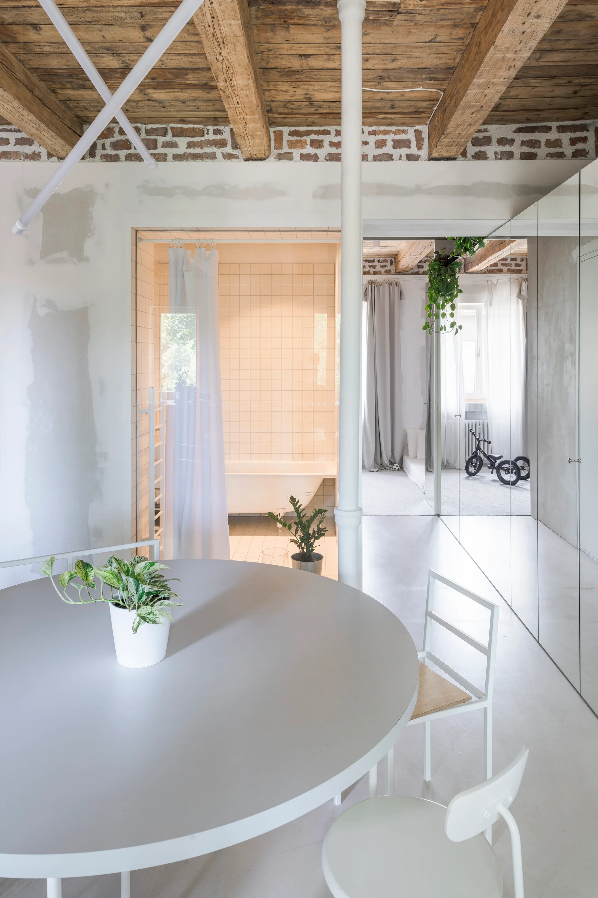 A minimalist round dining table and white chairs in a modern loft space featuring exposed ceiling beams and a glass-walled bathroom.