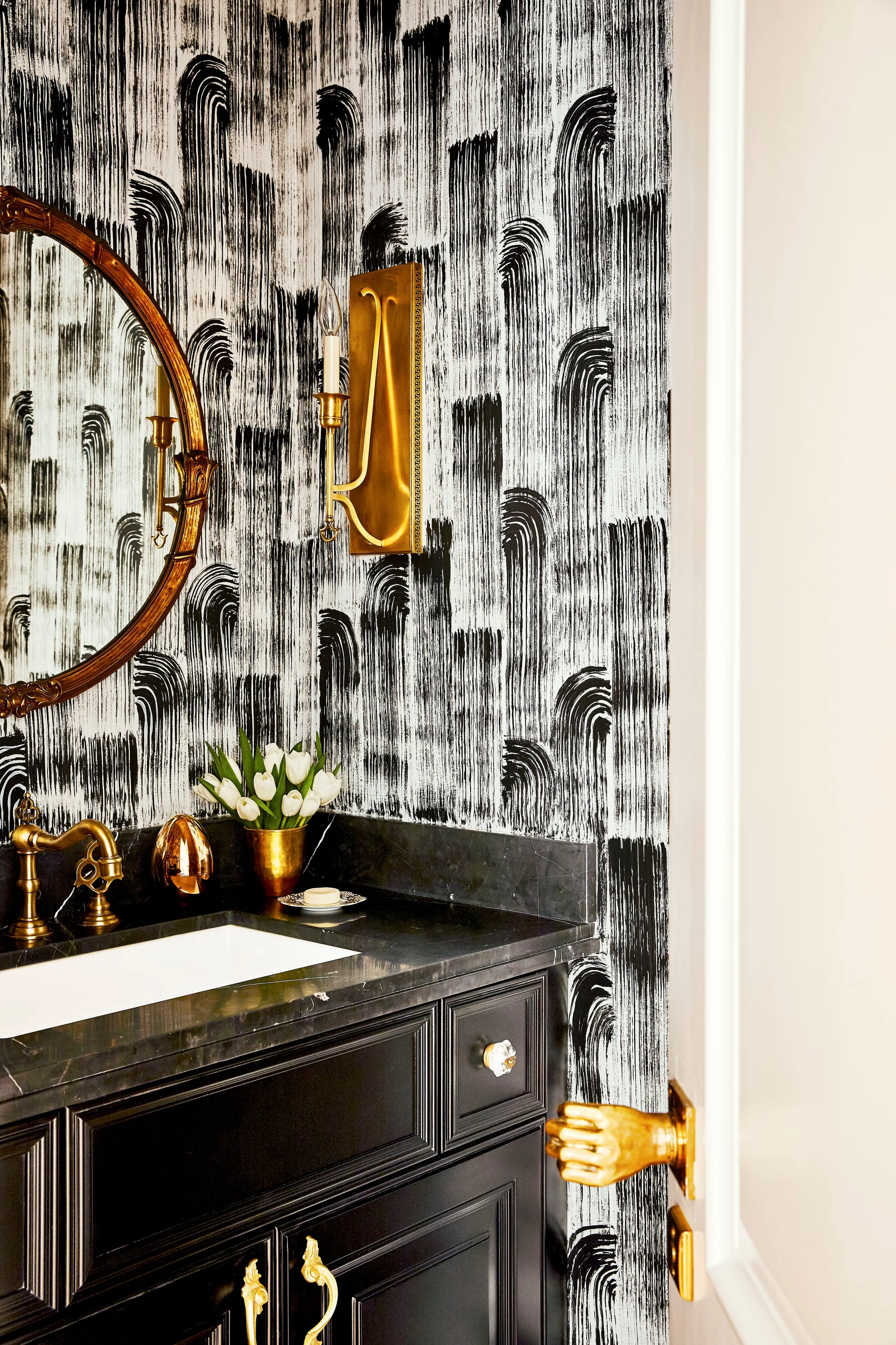 A powder room features black and white brushstroke wallpaper, a dark stone vanity with a brass faucet, and an ornate brass sconce.