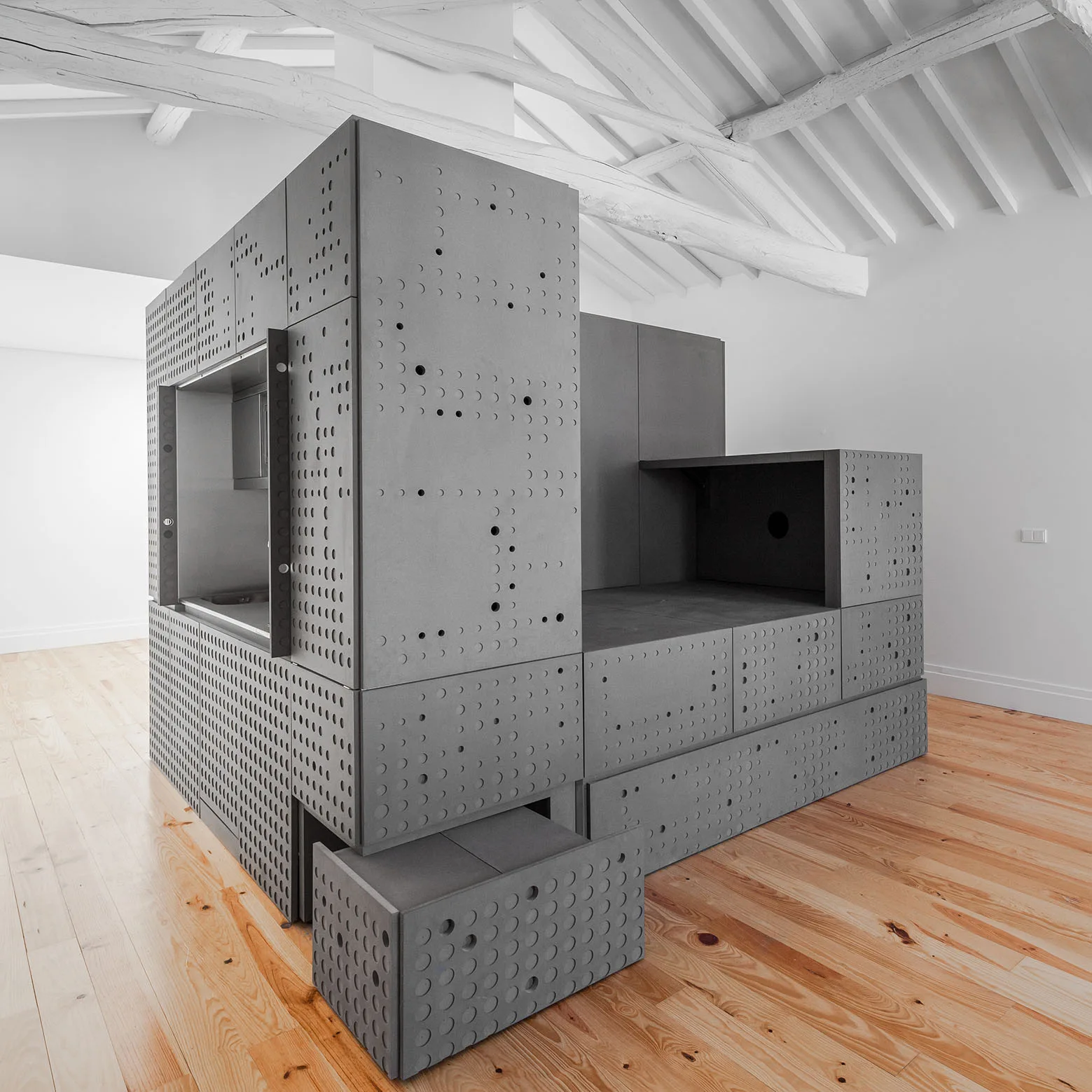 A large grey modular storage unit with perforated circular holes sits on a light wood floor beneath white painted ceiling beams.